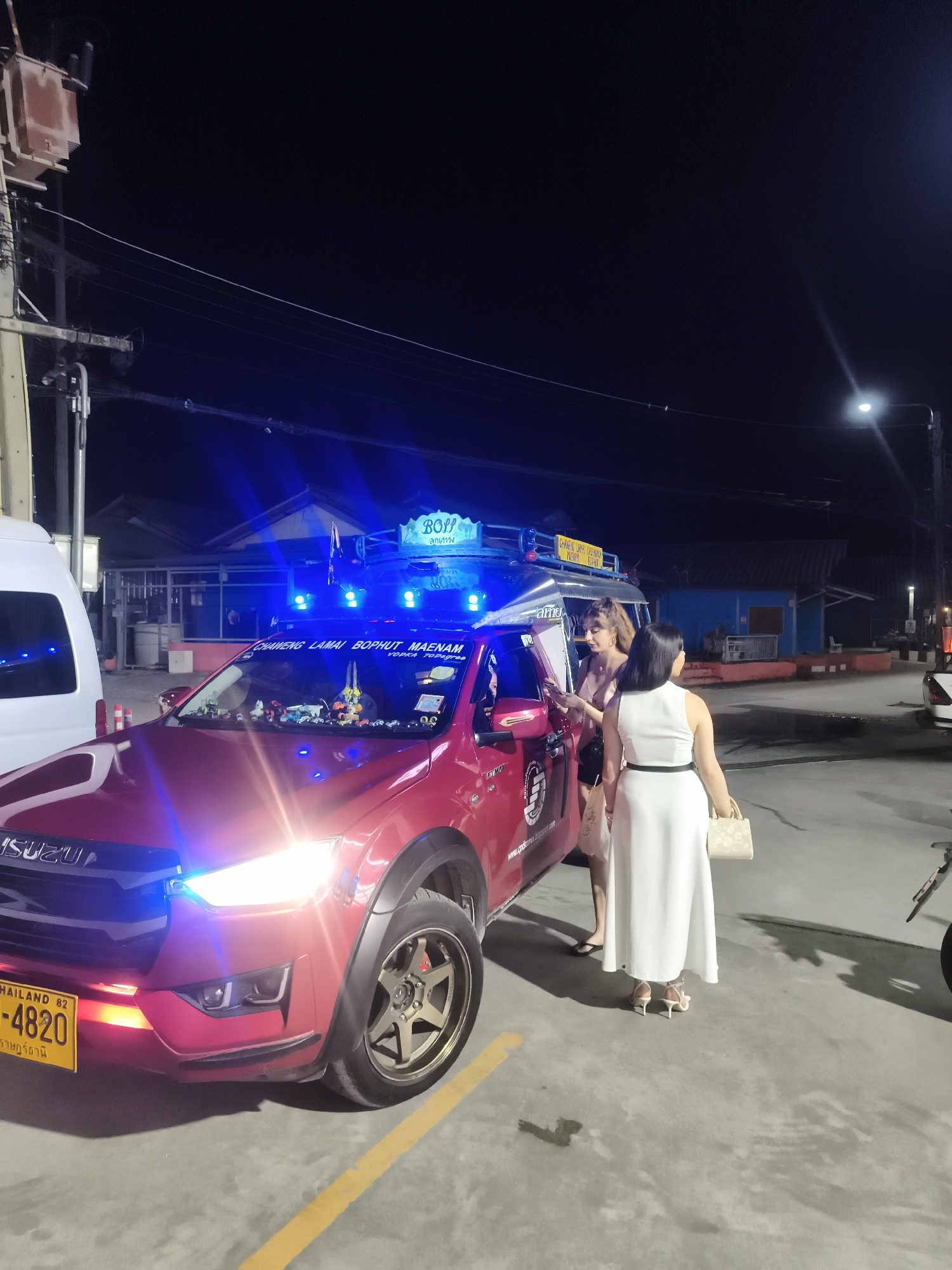 Here is a concise description of the image:
At night in Baan Chaweng, Thailand, a red Isuzu truck with numerous blue lights and rooftop signage reading "Boss" is parked on a paved surface marked with yellow lines. Two women are standing near the truck. One woman is wearing a white dress and carrying a light-colored purse. The other woman is wearing a short, pink outfit. The truck’s license plate reads "THAILAND 82 P-4820." The environment includes buildings, street lights, and power lines.