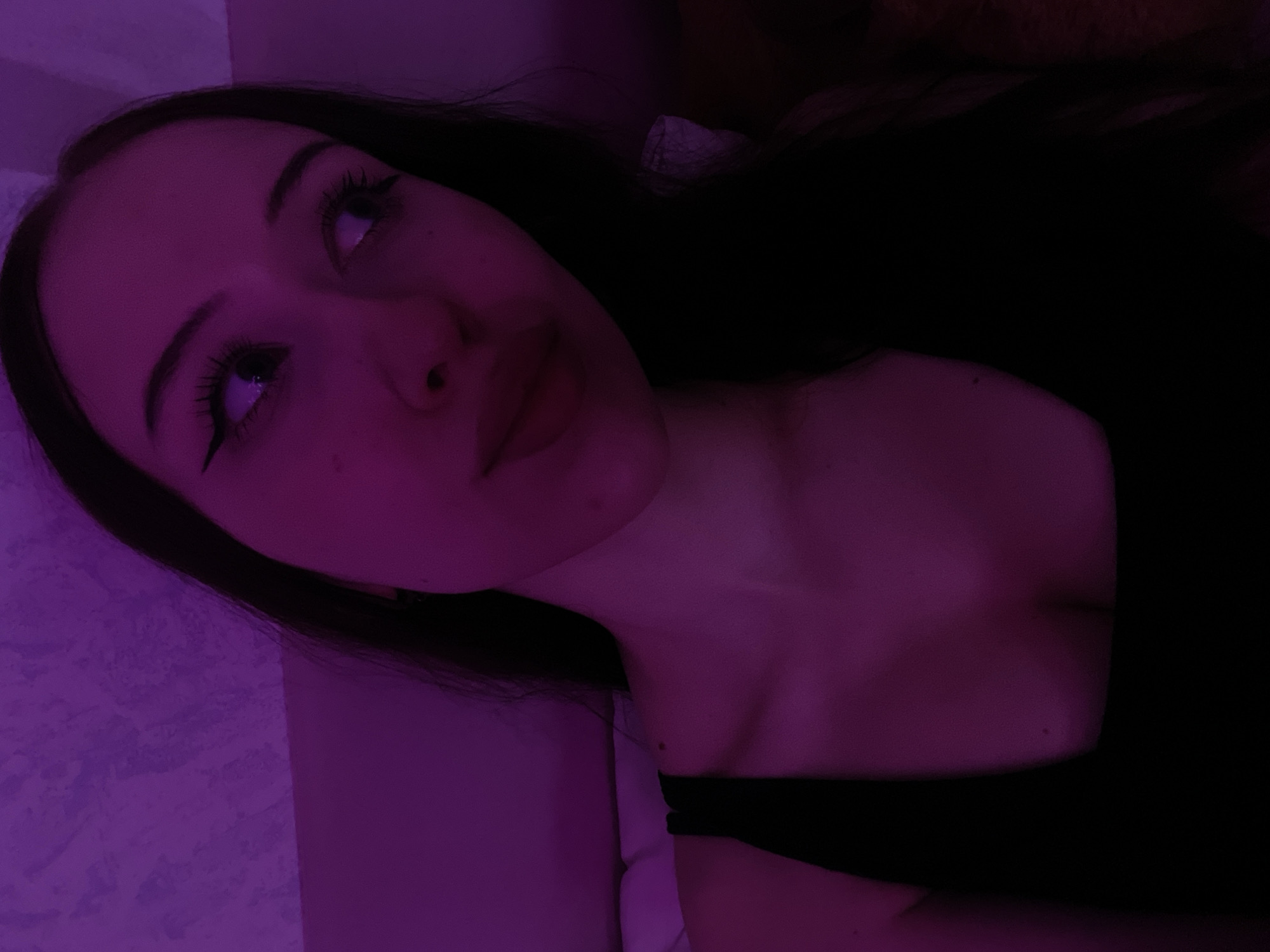 A young woman with long dark hair, wearing a black top, is pictured in a dimly lit room. The lighting casts a purple hue across her face and shoulders. She has winged eyeliner and is looking off to the right. The background appears to be a textured wall. There is no indication of the location being in Votkinsk, Russia, or any other specific city.