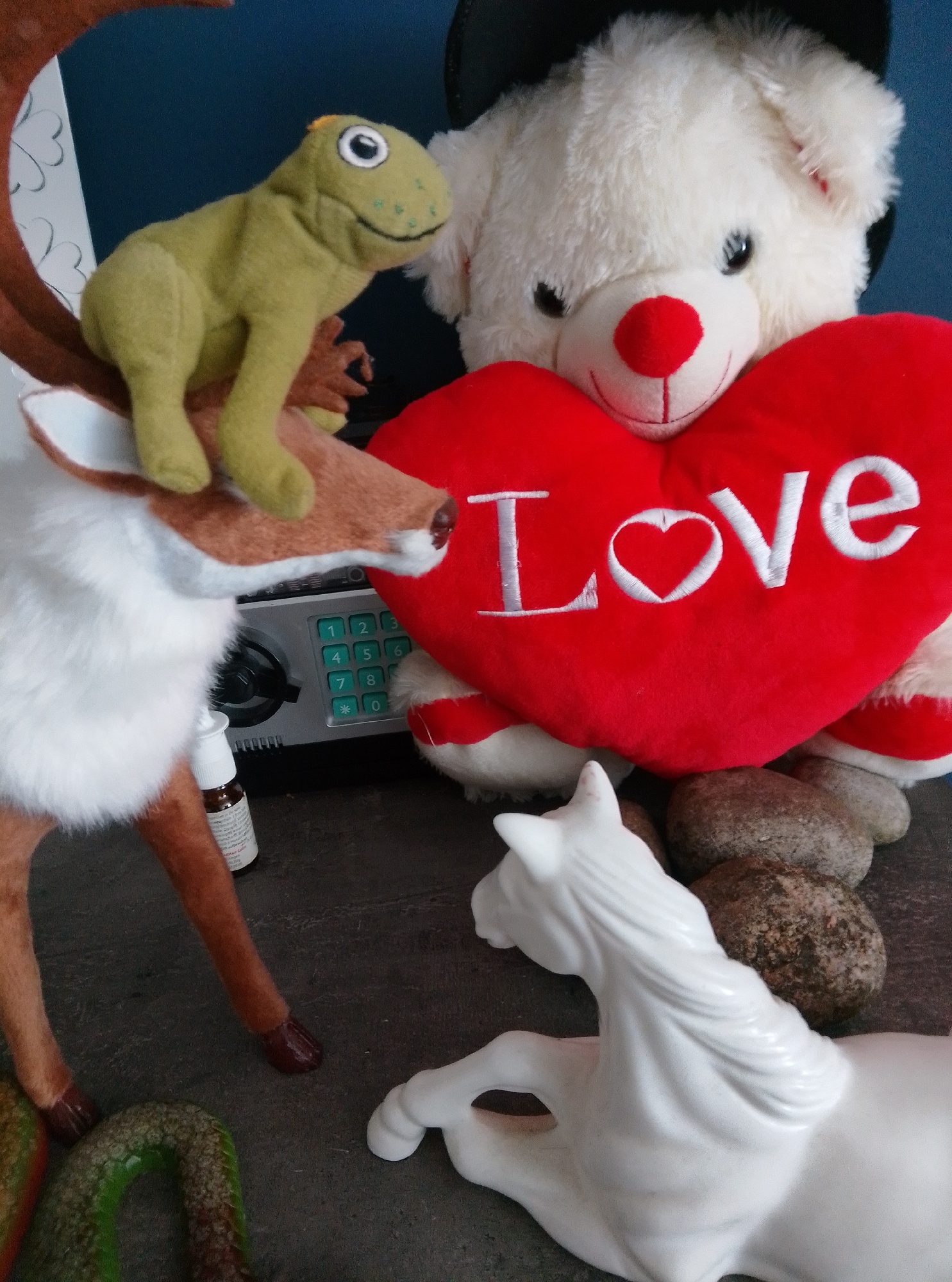 A collection of stuffed animals and decorative objects are arranged on a surface. In the foreground, a white ceramic horse is positioned on the right. Behind it, a cluster of rounded, grey stones are visible. To the left of the horse, a plush reindeer with brown antlers and white fur is partially visible. A green plush frog is perched on the reindeer's antlers. A red heart-shaped cushion with "Love" embroidered in white lettering is placed in the center, with a white teddy bear partially obscuring it. A small bottle with a white cap is situated near the reindeer's legs. Behind the reindeer and to the left of the heart cushion, a grey metal safe with a green numerical keypad is visible. The background consists of a dark blue surface. The scene does not contain any identifiable information to determine its location in the city of Wedel, Germany.