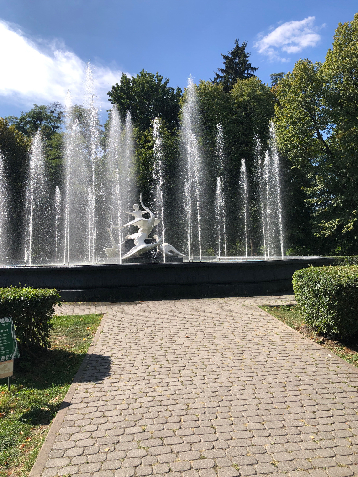 A fountain with a sculpture of figures in motion is the central subject of this image, located in a park in Lviv, Ukraine. The fountain features multiple water jets creating a dynamic display against a backdrop of lush green trees and a blue sky with scattered clouds. A paved walkway leads towards the fountain, bordered by neatly trimmed bushes and grass. The scene appears to be a public recreational area, likely in a park or garden setting within the city.