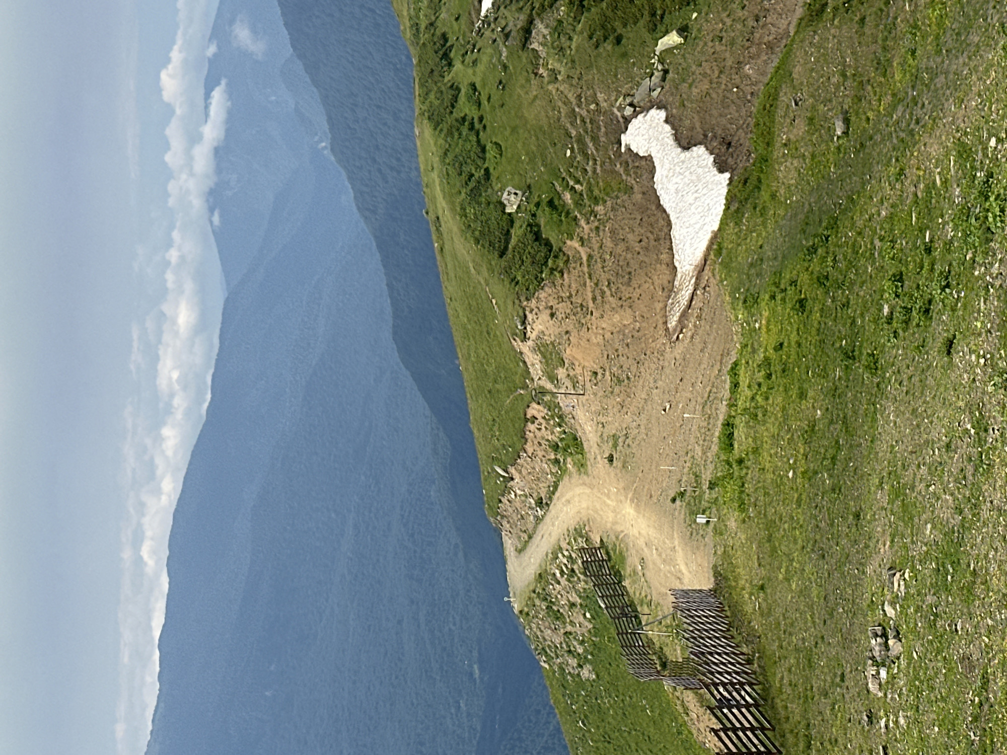 An overhead, medium shot captures a scenic view of a dirt path winding up a grassy mountain slope. The path is bordered by a wooden fence on the left side of the image, constructed from horizontal planks supported by vertical posts. The fence guides the path, which curves gently as it ascends. The mountain slope is covered in vibrant green grass, with patches of exposed dirt and rocks scattered throughout. Further up the slope, a small patch of snow can be seen.
In the background, a series of majestic mountains stretch across the horizon, their peaks fading into a hazy blue due to atmospheric perspective. The sky above is overcast, with soft, white clouds adding a gentle texture to the scene. The overall impression is one of natural beauty and rugged terrain, hinting at a remote and elevated location.