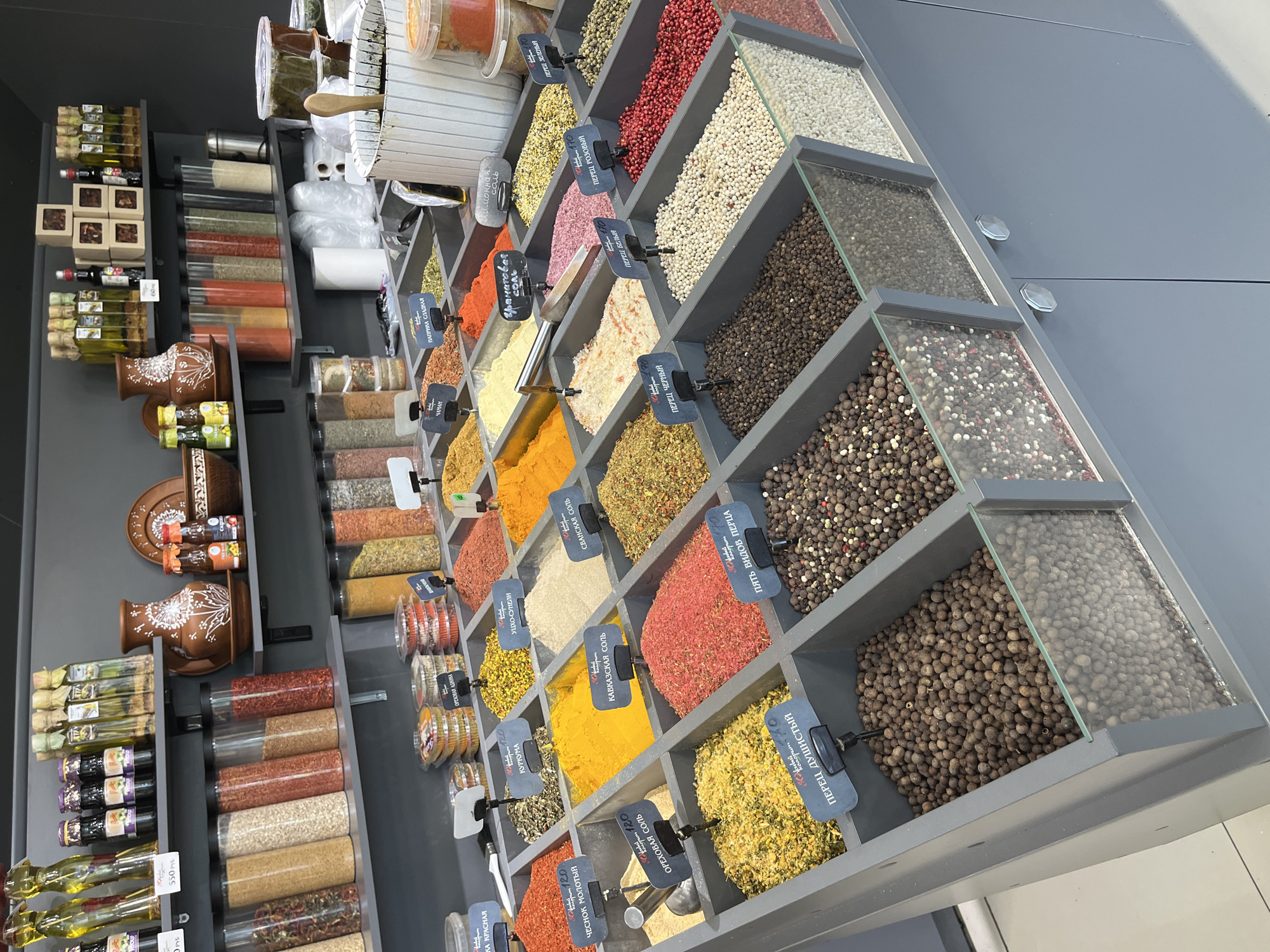 This is a medium shot, angled photograph of a display of spices in what appears to be a market or store. The spices are organized in a tiered shelving unit with individual compartments, many of which have clear glass fronts to showcase the contents. The majority of the compartments are filled with a variety of dried spices in different colors, textures, and forms, including whole peppercorns, ground powders, and flakes. Several small black labels with white text are attached to the front of the compartments, identifying the spices in what appears to be Cyrillic script. Visible text includes "ЧЕСНОК МОЛОТЫЙ," "КУРКУМА," "ОРЕХОВАЯ АДЖИКА," "СВАНСКАЯ СОЛЬ," "ПЕРЕЦ ЧЕРНЫЙ," and "ПЯТЬ ВИДОВ ПЕРЦА."
In addition to the spices, there are also bottles of oil and other liquids on lower shelves, as well as some decorative ceramic pottery. The lighting suggests an indoor setting, possibly during the daytime, with no indication of weather conditions or specific time of day. The overall impression is one of abundance and variety, focusing on the colorful and diverse nature of the spices. There are no people visible in the image.