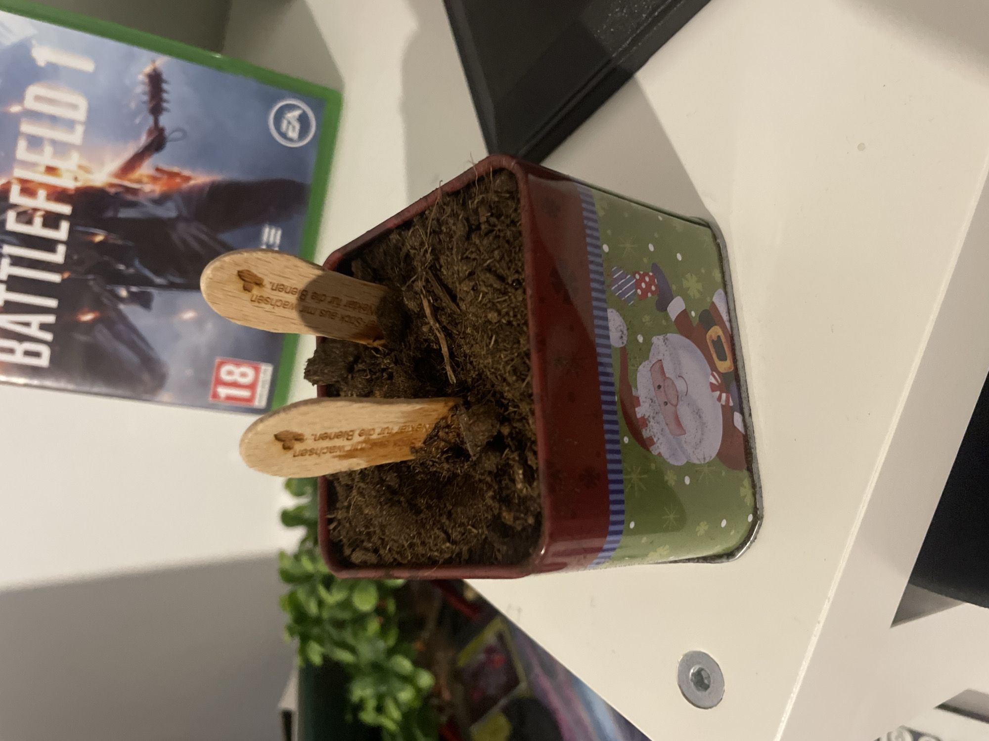 The image shows a square, festive-themed planter filled with soil and two small wooden labels with text in German, sitting on a white surface. A video game case titled "Battlefield 1" is visible in the background. A green plant is partially visible at the bottom of the image. The scene appears to be indoors, on a shelf or similar surface. The specific location is not determinable from the provided visual information.