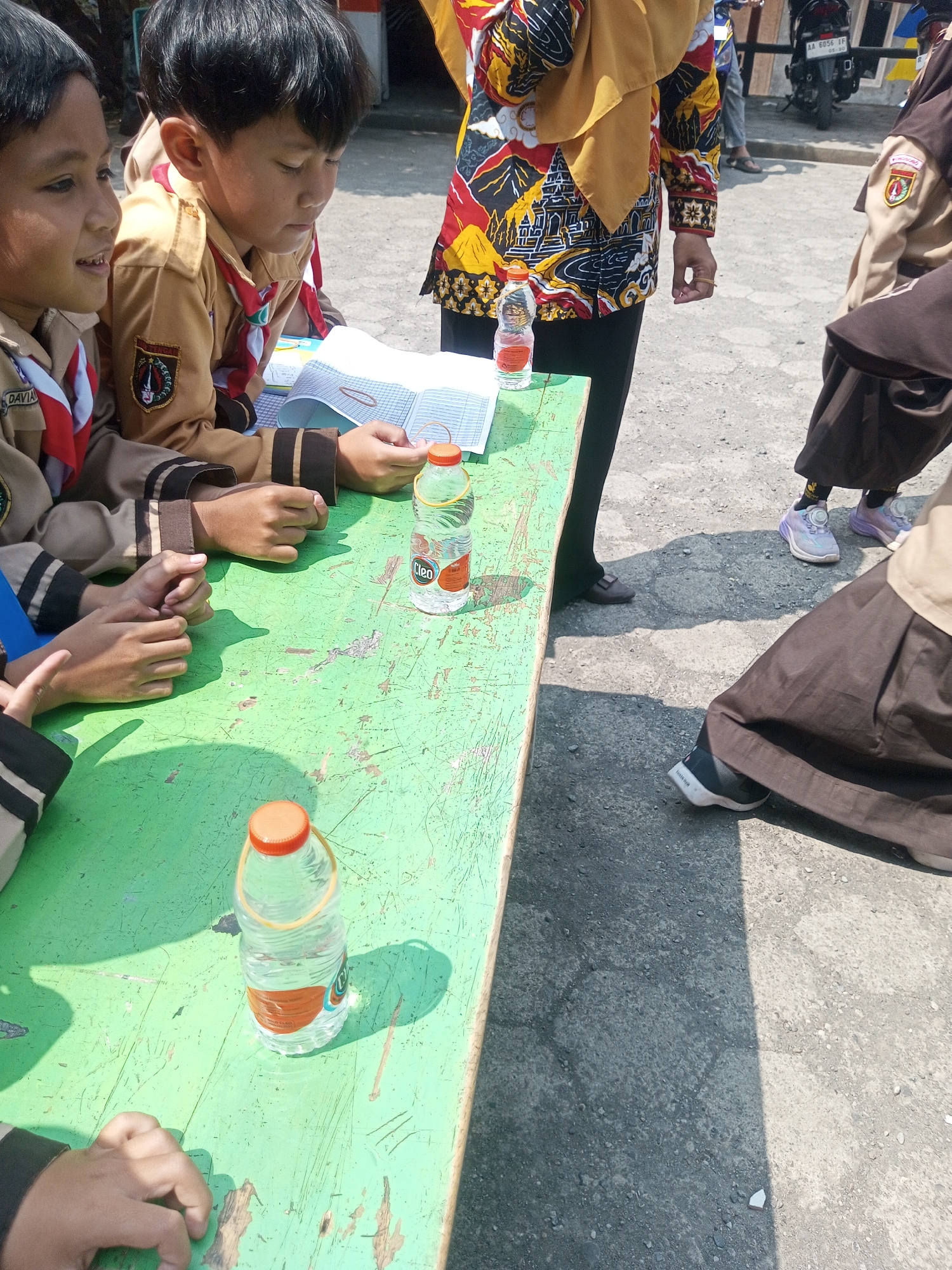 The image depicts a group of children, likely students, gathered around a weathered, green wooden table outdoors, possibly in the grounds of a school in Kalibebe, Indonesia. The children are wearing uniforms, which appear to be light brown shirts with darker trim and red scarves. Two boys are seated at the table, looking down at some papers. One boy is wearing a light brown shirt with a patch on the sleeve. Another boy is wearing a similar uniform. Several plastic water bottles with orange caps are on the table. In the background, other individuals are visible, including one wearing a colorful patterned shirt and dark pants, and others in dark skirts and light-colored shoes. A motorcycle is parked behind them. The ground is a rough, gray asphalt or concrete surface. The lighting suggests it is daytime, with strong sunlight casting shadows.
