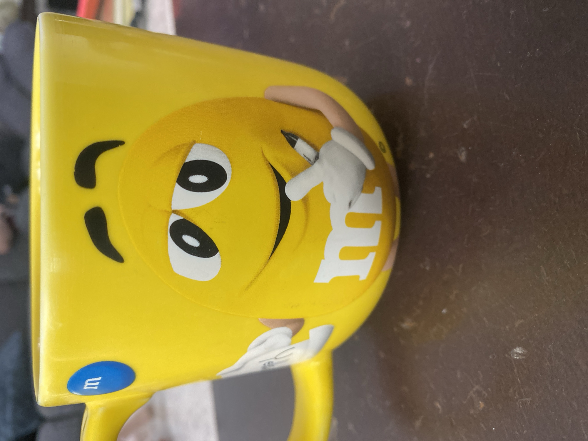 The image displays a vibrant yellow M&M's character ceramic mug, presented horizontally on a dark brown, somewhat textured surface. The mug features a large, smiling yellow M&M character with expressive eyes and a wide, cheerful grin. The character's left hand, depicted in a white glove, holds a small black marker or pen, poised as if about to write. The distinctive white 'm' logo is clearly visible on the character's body. On the mug's handle, there is a smaller blue M&M emblem, also adorned with a white 'm'. The background is blurred, indicating an indoor setting with diffused lighting. The dark surface beneath the mug shows minor imperfections like scuffs and small specks of dust. The overall scene captures the playful design of the mug in a static, indoor shot from San Bernardino Tlaxcalancingo, Mexico.