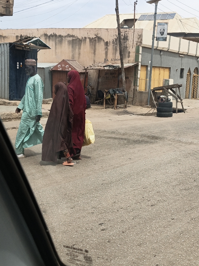The scene is a daylight street view in Hotoro South, Nigeria, captured from within a vehicle.
In the foreground, a paved asphalt road, light grey with visible aggregate, extends across the frame. Three individuals are walking from left to right. The first, an older male with a white beard, wears a light blue traditional garment and a patterned cap. He is mid-stride, with his left foot in a white slipper visible. Following him is a smaller individual, likely a child, wearing a dark brown full-length garment over patterned leggings and orange sandals. Behind them, an adult female in a dark red full-length garment carries a yellow plastic bag in her right hand; only her feet in dark sandals are visible.
The midground features a collection of commercial structures and buildings. On the left, a dark blue corrugated metal kiosk with a green canopy bears the faded inscription "FORENSIC VENTURE." To its right, a weathered, light brown wall with stains and graffiti ("BA ADAWU") spans the background. Further right, a small brown corrugated metal stall is visible, followed by a wooden makeshift stall or workbench with various items. Behind this stall, faded writing on a wall reads "ZA BATI DIRI MU." To the far right, a grey building with a golden-yellow corrugated metal shutter on the ground floor and a decorative golden-yellow metal gate is visible. The building has a light-colored, possibly tiled, pitched roof.
A wooden utility pole stands on the right, supporting overhead power lines and a metal pole. Attached to the metal pole is a solar panel at the top and a banner below it, displaying "TARAYA TI" and a portrait of a man with additional text (partially visible "DR ALIYU..."). Near the base of the utility pole, three black tires are stacked horizontally, next to which a small internal combustion engine (possibly a generator) rests on a stand. The sky is hazy light blue.