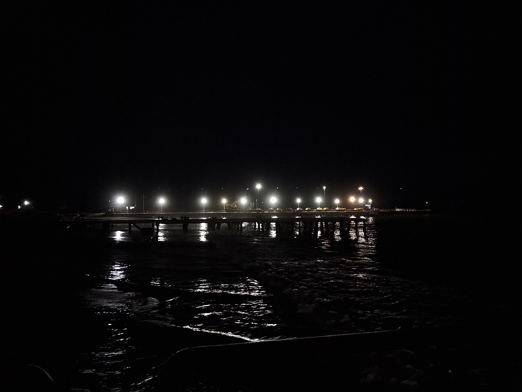 The image depicts a nighttime scene at the pier in Chimbote, Peru. A long, illuminated pier stretches across the frame, with a series of bright lights lining its edge. The lights cast reflections onto the dark water below, creating shimmering patterns. The water appears choppy, with some foamy waves near the foreground. The sky is completely dark, indicating it is nighttime. There are no visible people or discernible objects on the pier or in the water, apart from the pier structure itself and the lights. The overall impression is one of quiet and solitude.