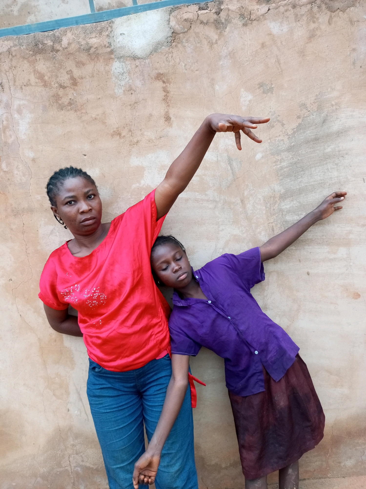 Two females, an adult and a child, stand in front of a textured, light brown wall, likely in Tempure, Nigeria. The adult, on the left, wears a bright red shirt and blue denim jeans, her left arm resting against the wall, and her right arm raised with fingers splayed. The child, to the right, wears a purple short-sleeved shirt and a dark red skirt, her head resting on the adult's shoulder and her eyes closed. The wall appears weathered and uneven, with visible imperfections and stains. The lighting suggests it is daytime.