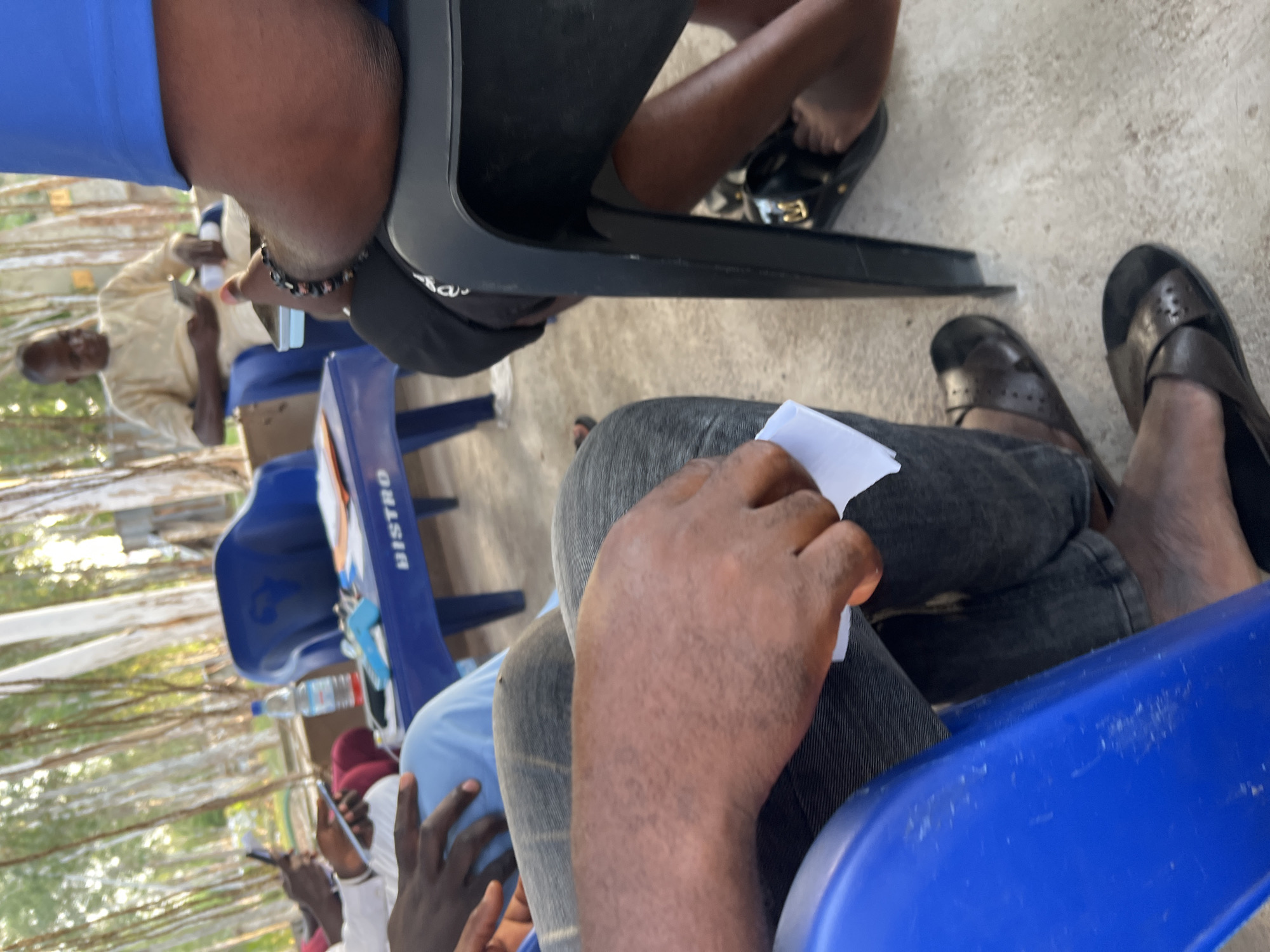 The image shows a group of people seated outdoors, likely in a casual setting. The subjects are primarily visible from the waist down, with some hands and arms also in frame. One person in the foreground is holding a small piece of white paper. Another individual is wearing a light-colored traditional garment and appears to be holding a phone. Several people are wearing dark-colored sandals or flip-flops.
The seating appears to be on blue plastic chairs, some of which have the word "BISTRO" printed vertically in white text on them. A partially visible bottle of water is also on one of the chairs. The ground is a light-colored concrete or paved surface. In the background, there are thin, vertical structures that resemble trees or reeds, suggesting an outdoor, possibly natural or semi-natural environment.
The lighting suggests it is daytime, with bright sunlight filtering through the trees in the background. No specific emotions are discernible from the cropped view. The overall impression is one of a relaxed gathering or meeting. The location context provided is Mayo Dasa, Nigeria, which fits the visual cues of an outdoor setting.