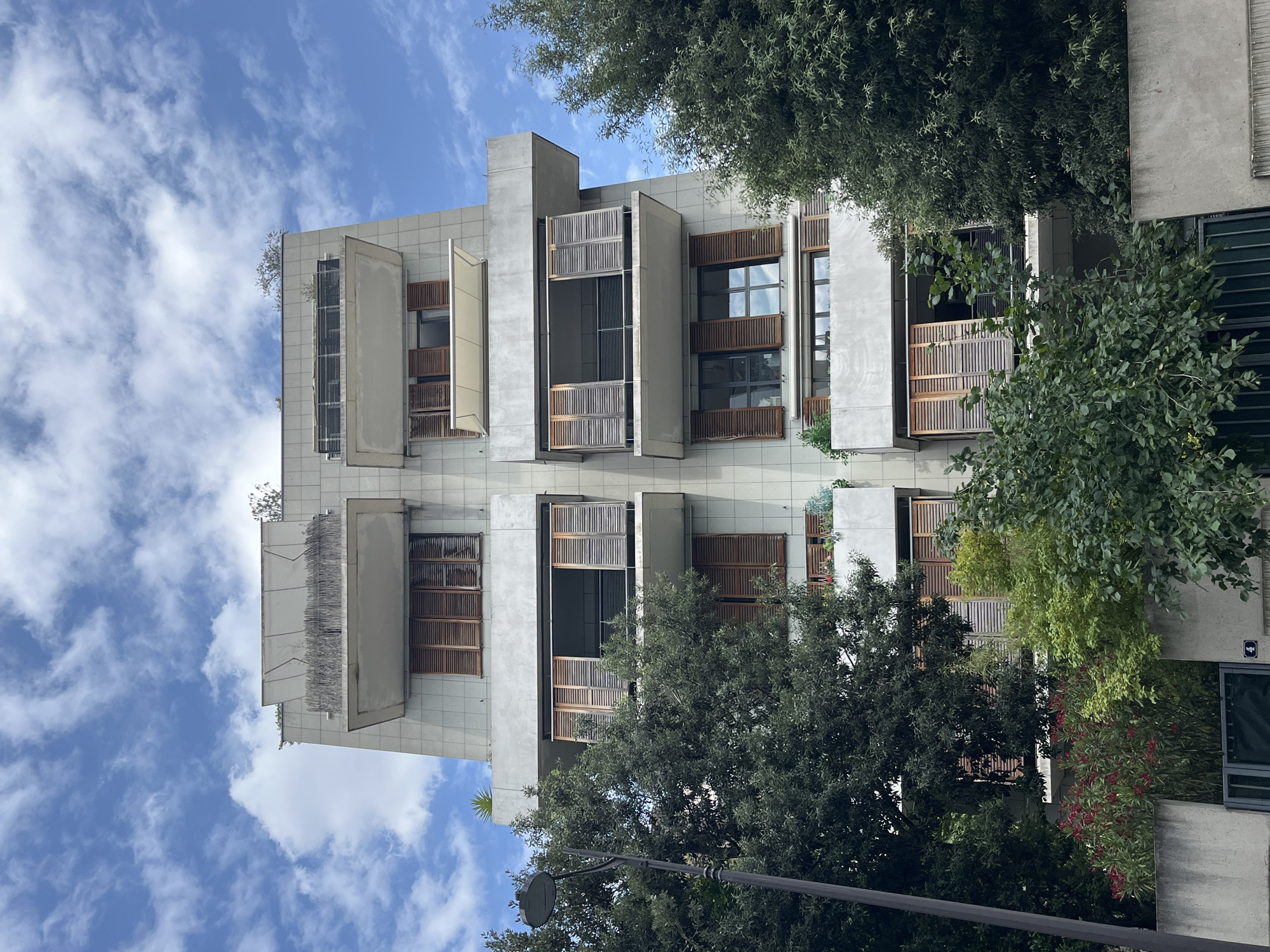 This is a vertical-oriented, eye-level shot of a modern building's facade, interspersed with lush greenery. The building features a contemporary design with concrete elements and large windows. Many of the windows are covered by louvered wooden shutters, some of which are open, revealing the darker interiors. Several windows also have attached planters, overflowing with foliage, adding a natural element to the architecture. The building's exterior appears to be clad in light-colored tiles.
The sky above is a vibrant blue, dotted with scattered white, wispy clouds, suggesting a bright and pleasant day. The time of day is likely daytime, given the natural lighting. The foliage is dense and varied, with some trees reaching up to frame the upper portions of the building and others cascading down the sides. This suggests an urban or suburban setting where nature is integrated into the built environment.
There are no people visible in the image. The focus is solely on the interplay between the building's architecture and the surrounding plant life, under a clear sky. The overall impression is one of a stylish, possibly residential or commercial building designed with a keen aesthetic sense and a connection to nature. No visible text is present in the image.