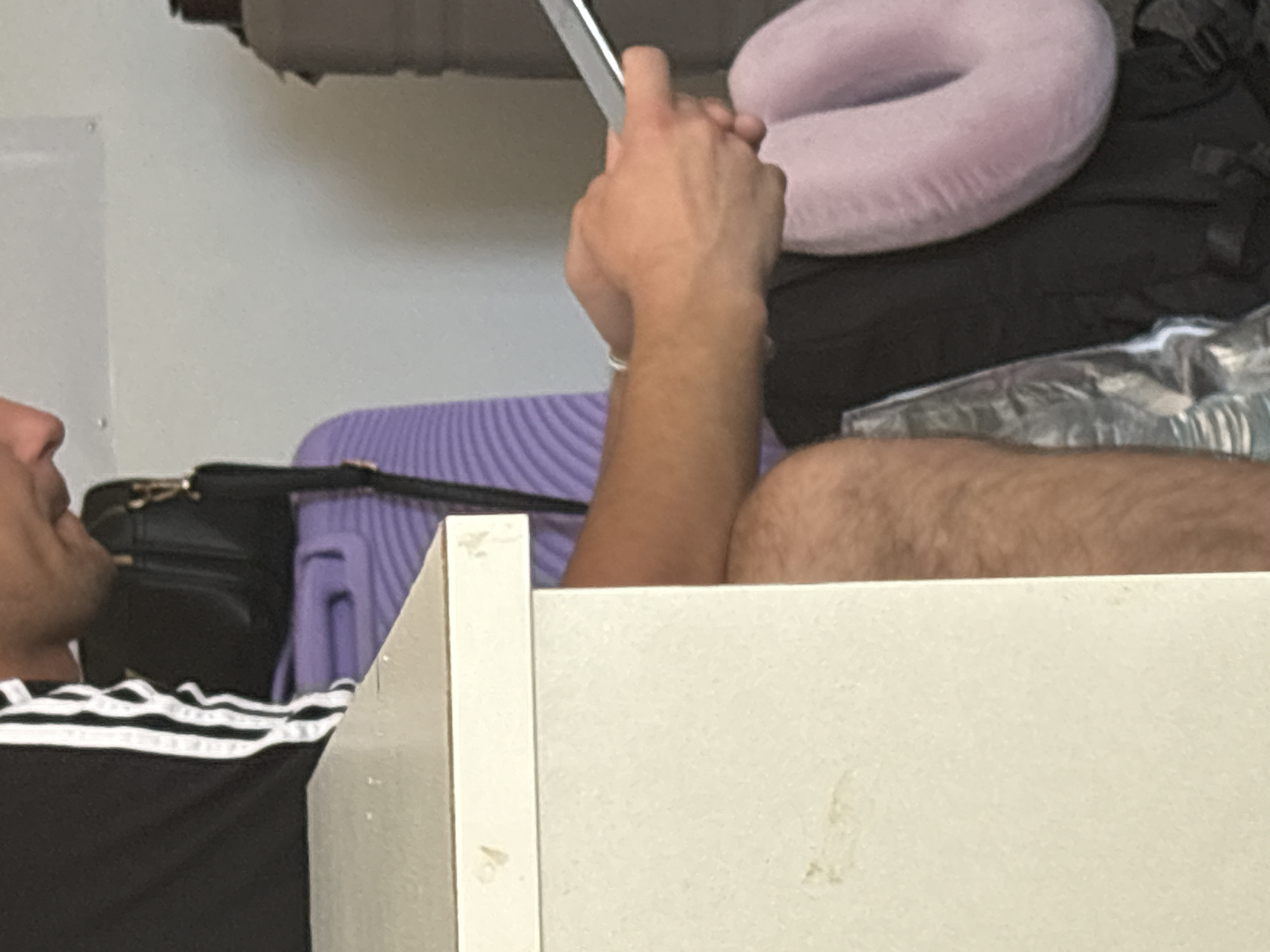 The image shows a close-up view of a person lying down, possibly in a room or on a trip. The person is wearing a black t-shirt with white stripes on the sleeves. Their arm, resting on their leg, is visible and appears to be hairy. In the background, there are several objects, including a purple suitcase, a black bag, a light pink travel pillow, and some other luggage. The person is holding a silver tablet or phone. The lighting suggests it might be daytime, but there are no strong indications of the time of day or weather. The setting appears to be an indoor space with a lot of luggage, which could imply travel or a temporary living situation. There is no visible text in the image.