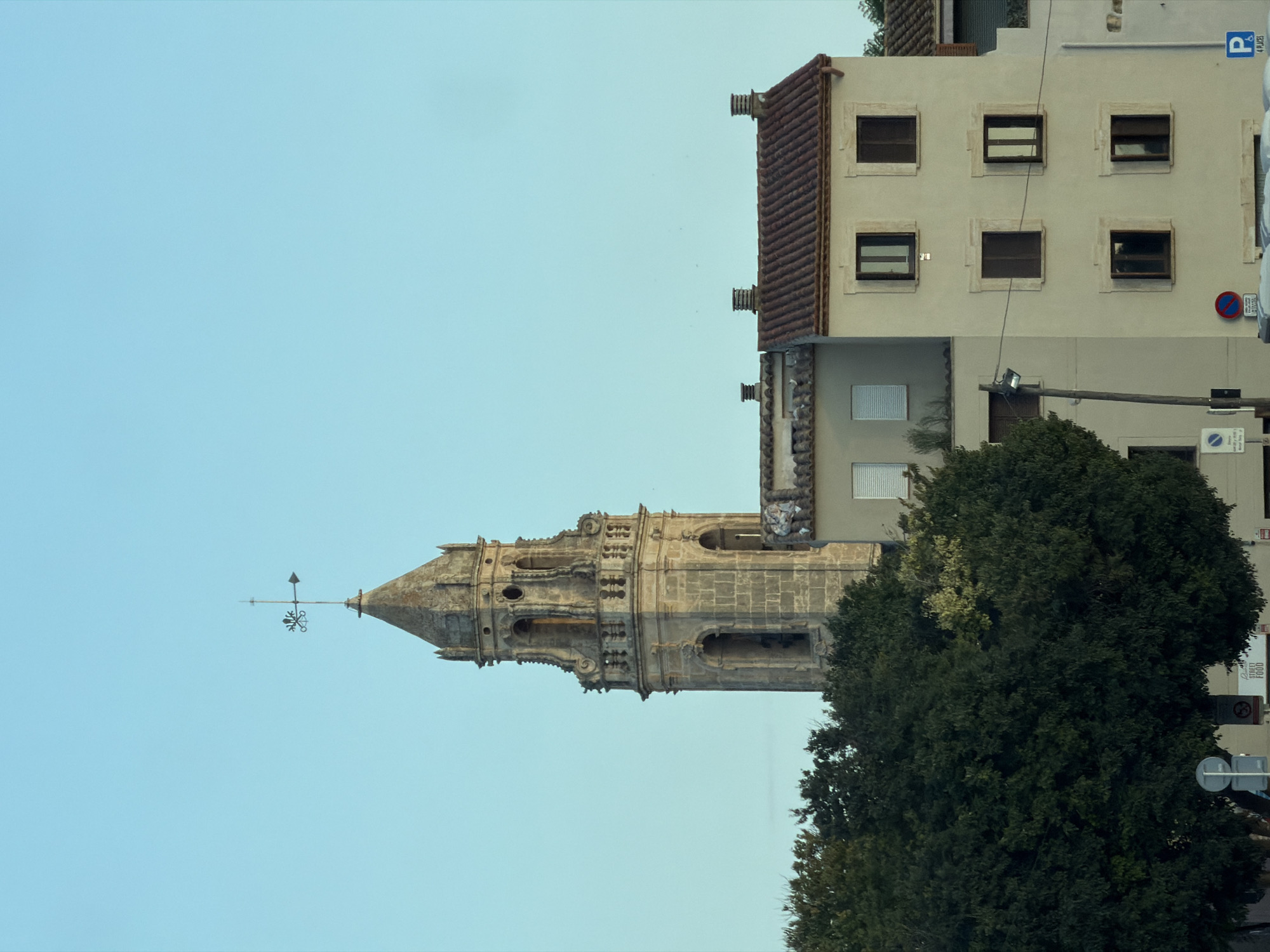 The image shows a view of buildings and a church tower in Torredembarra, Spain. In the background, a stone church tower with ornate detailing and a weathervane stands tall against a clear blue sky. In the foreground, a cluster of trees with dark green foliage partially obscures the lower section of the tower. To the right of the tower, a pale yellow building with a red-tiled roof is visible, featuring multiple windows. Another building, lighter in color and with horizontal white blinds on its windows, is situated to the left of the yellow building. The scene appears to be an outdoor urban landscape during daylight.
