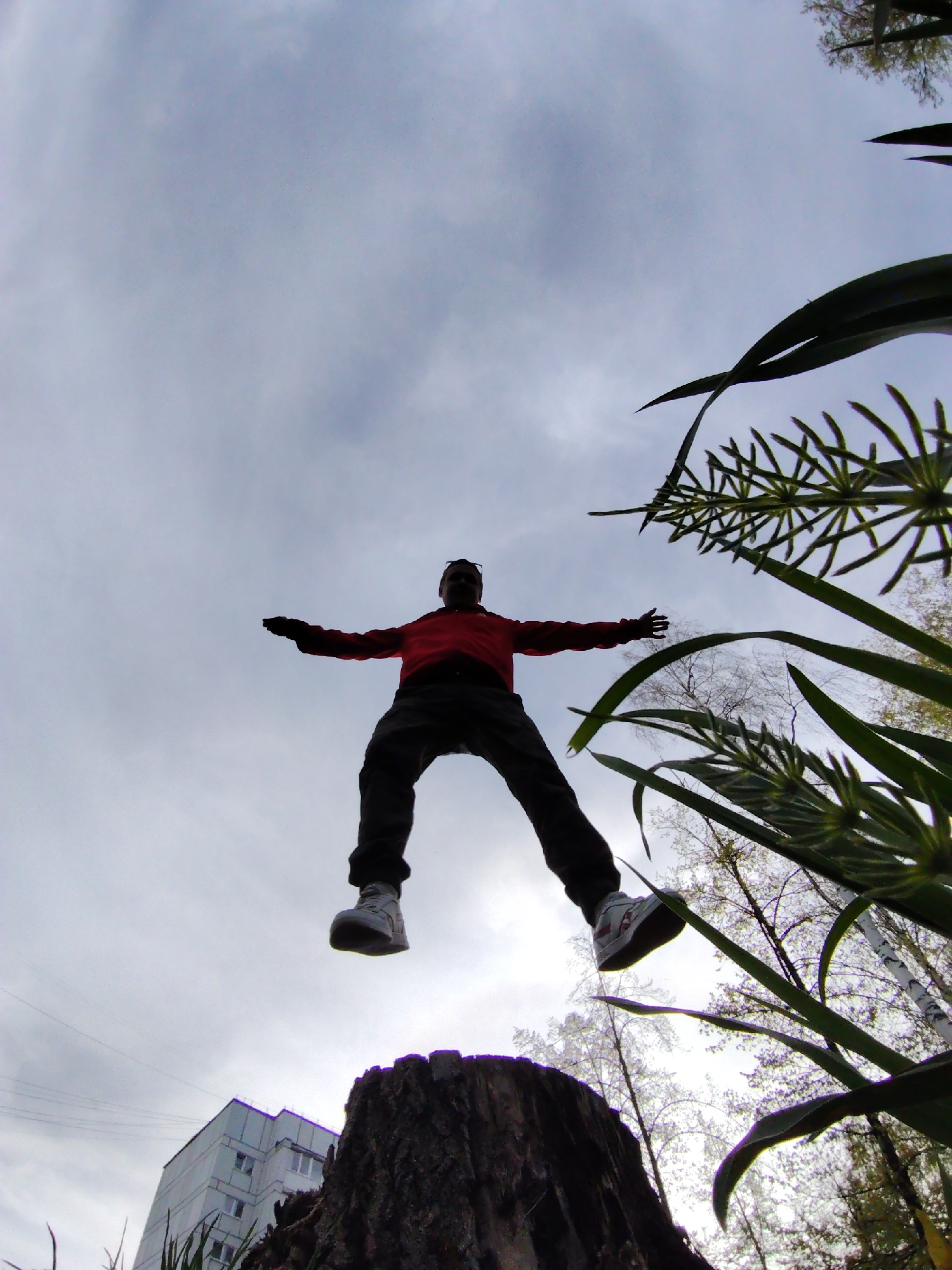 The image, taken from a low angle, depicts a person in mid-air, limbs outstretched, appearing to jump from a tree stump. The person wears a red jacket, dark pants, and white shoes. Green foliage fills the right side of the frame. A building is partially visible at the bottom left corner. Bare trees can be seen in the background. The sky is overcast with gray clouds. Given the reference to Chaikovsky, Russia, it can be inferred the image was taken there, although the specific location within the city is not identifiable from the visual information alone.
