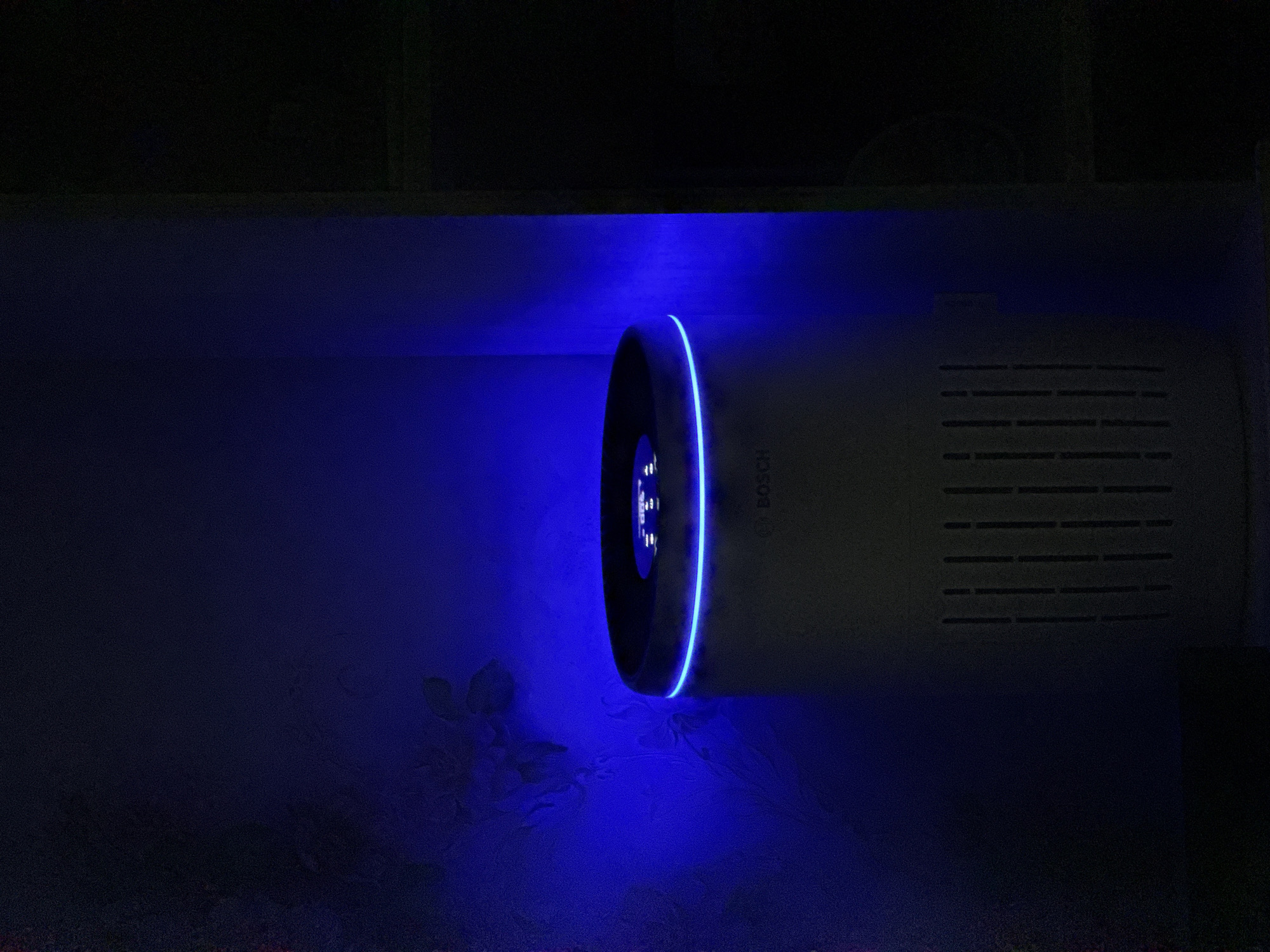 The image shows a white Bosch air purifier with a blue LED light ring around its top. The device is placed in a dark room, likely a residential interior, with a blue light illuminating the wall behind it. The wall appears to have a subtle floral pattern. The air purifier has a slatted vent on its lower front. The scene is not identifiable as being located in Slavyani, Bulgaria or any other specific city. There are no people or interactions depicted.