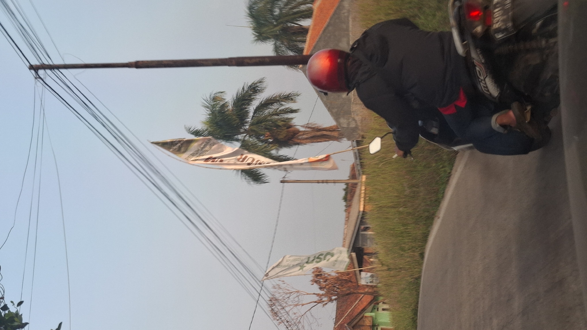 A person wearing a red helmet and a dark jacket rides a motorcycle on a paved road in Cibinong Hilir, Indonesia. The person is in the foreground, facing away from the camera. The motorcycle has a license plate that reads "F 5391 DB". There are two flags with text on them visible in the background, as well as some houses and palm trees. The sky is clear and blue. The overall lighting suggests it is either early morning or late afternoon.