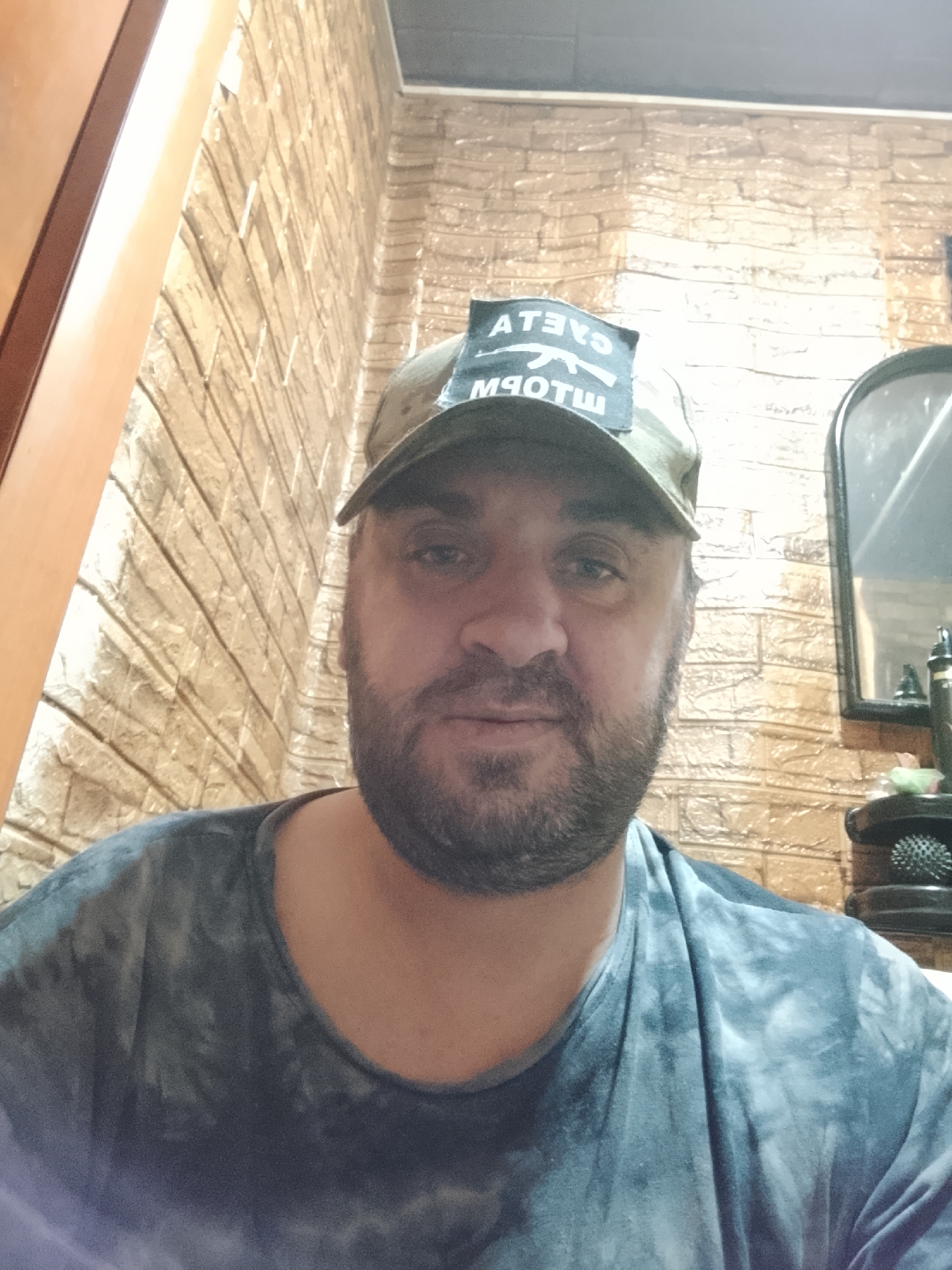 The image shows a medium shot of a man wearing a green baseball cap and a grey tie-dye t-shirt. The cap has a patch on the front with white text that appears to be reversed, and a silhouette of a rifle. The man has a beard and appears to be looking directly at the camera. The background consists of a brick-patterned wallpaper. To the right of the man, a portion of a mirror is visible, reflecting a dark object. The lighting is somewhat dim.