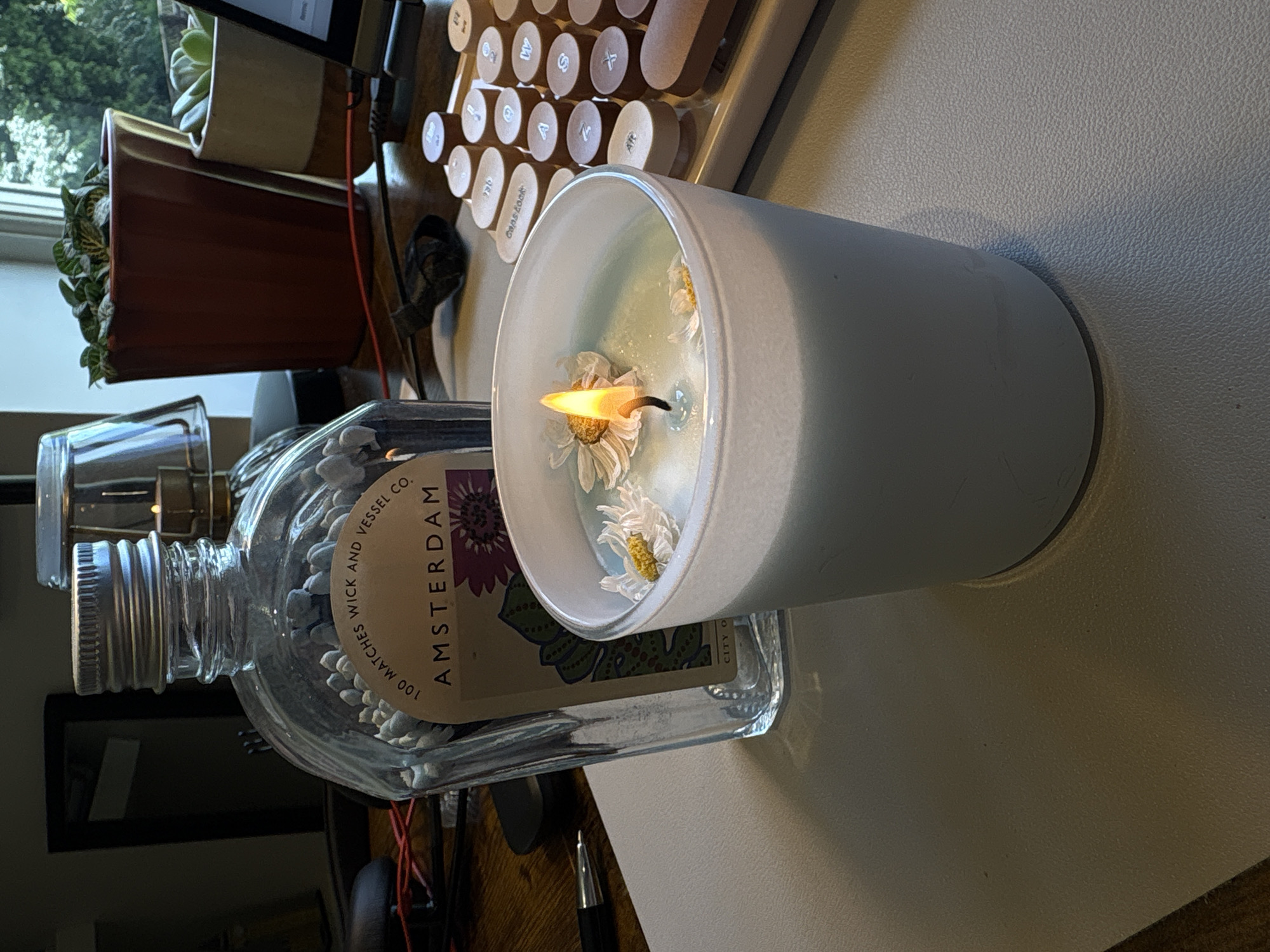 A lit candle with daisies floating in the wax sits on a desk next to a bottle of matches. The candle is in a white cylindrical container. The match bottle is clear glass with a silver lid and a label that reads "100 MATCHES WICK AND VESSEL CO. AMSTERDAM". In the background are plants in terracotta pots, a vintage-style keyboard, and what appears to be a desk or table surface. The scene is indoors. The location in the city of Ipswich, United Kingdom is unknown.
