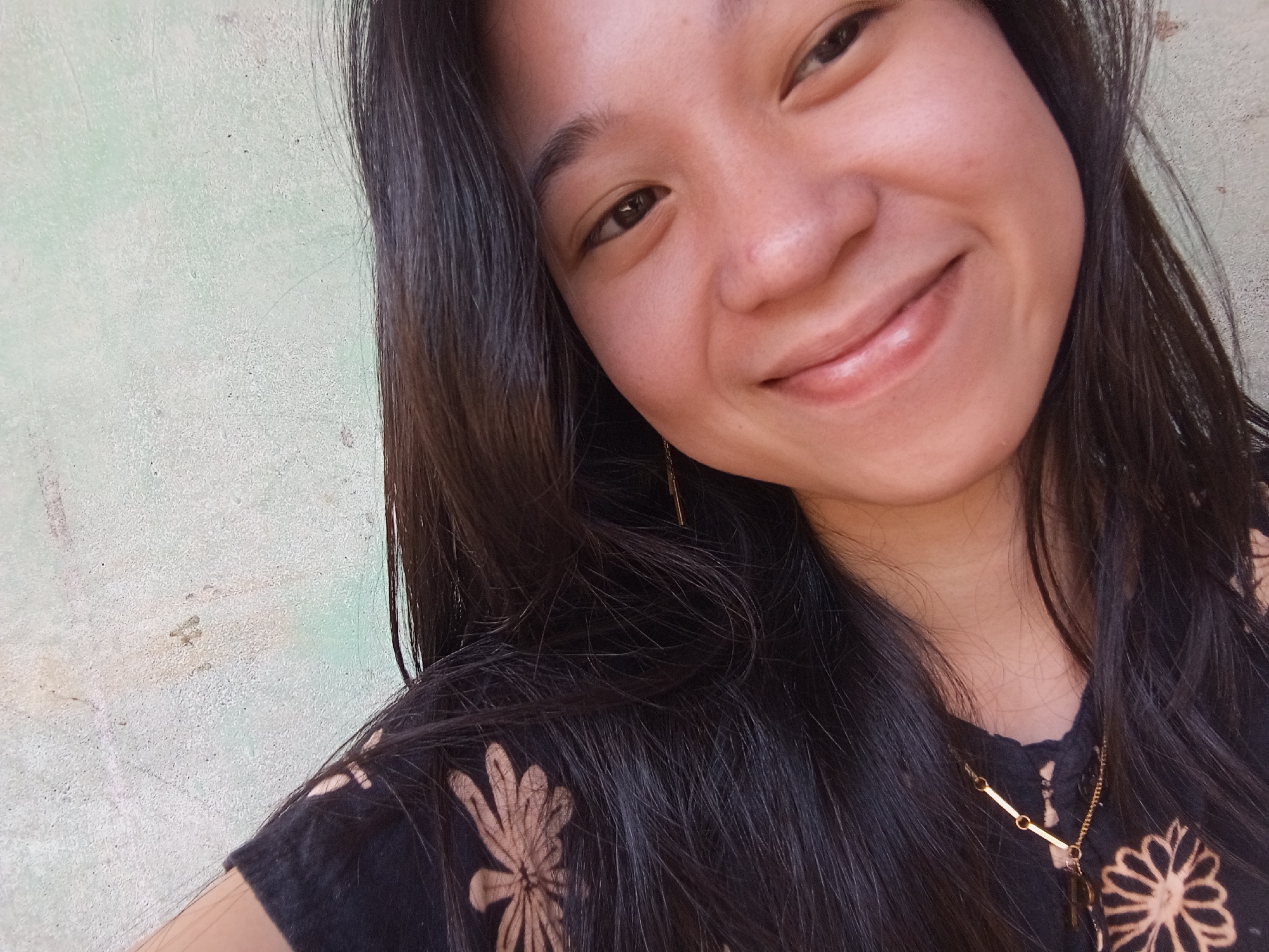 A young woman with dark hair smiles at the camera, her face angled slightly to the left. She wears a black shirt with a light-colored floral pattern and a delicate gold necklace. The background is a textured, light green wall. The image appears to be a selfie, taken outdoors in Rejosari, Indonesia.