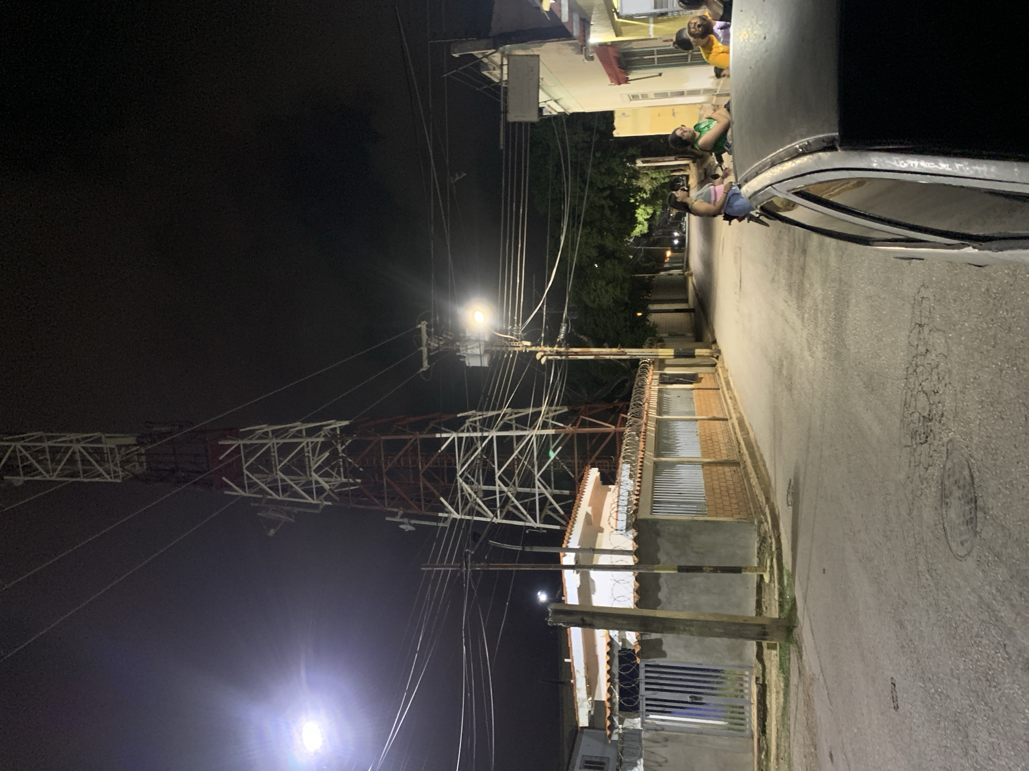 This image captures a nighttime street scene in Santa Ana, Venezuela. A prominent red and white lattice transmission tower dominates the upper left, extending into the dark sky, with numerous utility lines crisscrossing overhead, supported by several utility poles.
On the left side of the street, a white house with a terracotta-tiled roof is visible behind a concrete wall topped with barbed wire, featuring a dark blue metal gate. The asphalt road in the foreground shows surface wear and patches. Streetlights mounted on utility poles illuminate the scene, casting bright light on the foreground and further down the street, where trees line the roadside. One utility pole further down the street on the right displays an electrical transformer.
In the immediate right foreground, the roof and side window of a dark-colored car are visible. Beyond the car, a group of four individuals is gathered near the facade of a building. One woman, wearing a light-colored top and blue jeans, stands beside what appears to be a motorcycle. Adjacent to her, a person in a green top is seated or leaning. Two other individuals, one in a yellow shirt and another in a dark top, are partially visible further to the right, suggesting an informal social interaction. The buildings on the right side of the street have light-colored exteriors.