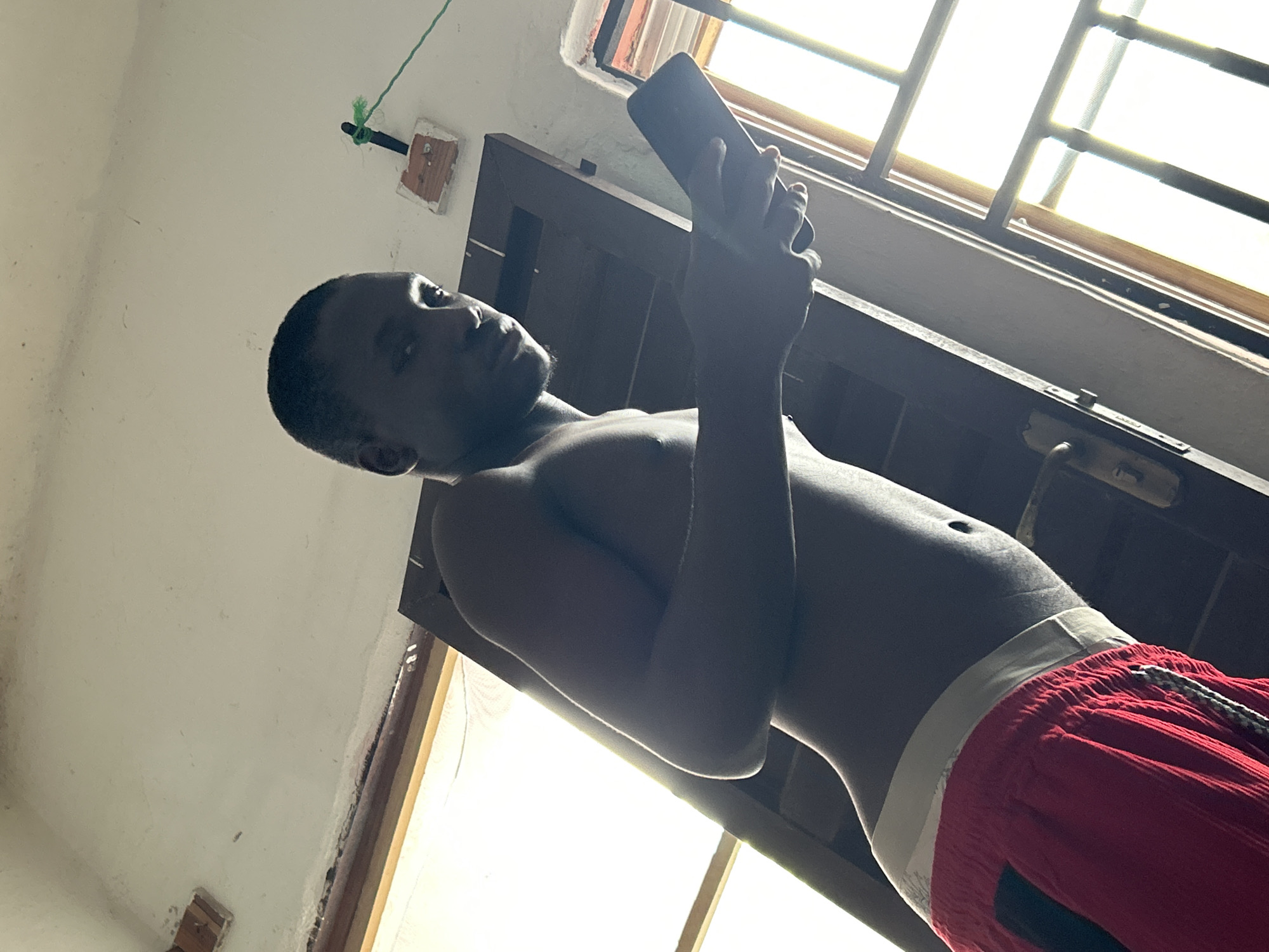 The image shows a young Black person, likely a male, standing shirtless indoors and looking down at a smartphone. The person is wearing red shorts with a white waistband and a drawstring. The background suggests an interior space with a window or skylight covered by bars, allowing bright natural light to enter. Part of a wooden doorframe is visible to the right. The lighting is strong, creating highlights and shadows on the subject's skin. The angle of the shot is from below, looking up towards the subject and the window. The scene appears casual, possibly a home environment. The time of day is suggested to be daytime due to the bright light. No text is visible in the image. The location context of Ihiagwa, Nigeria, aligns with the visual cues of the setting and the subject's appearance.