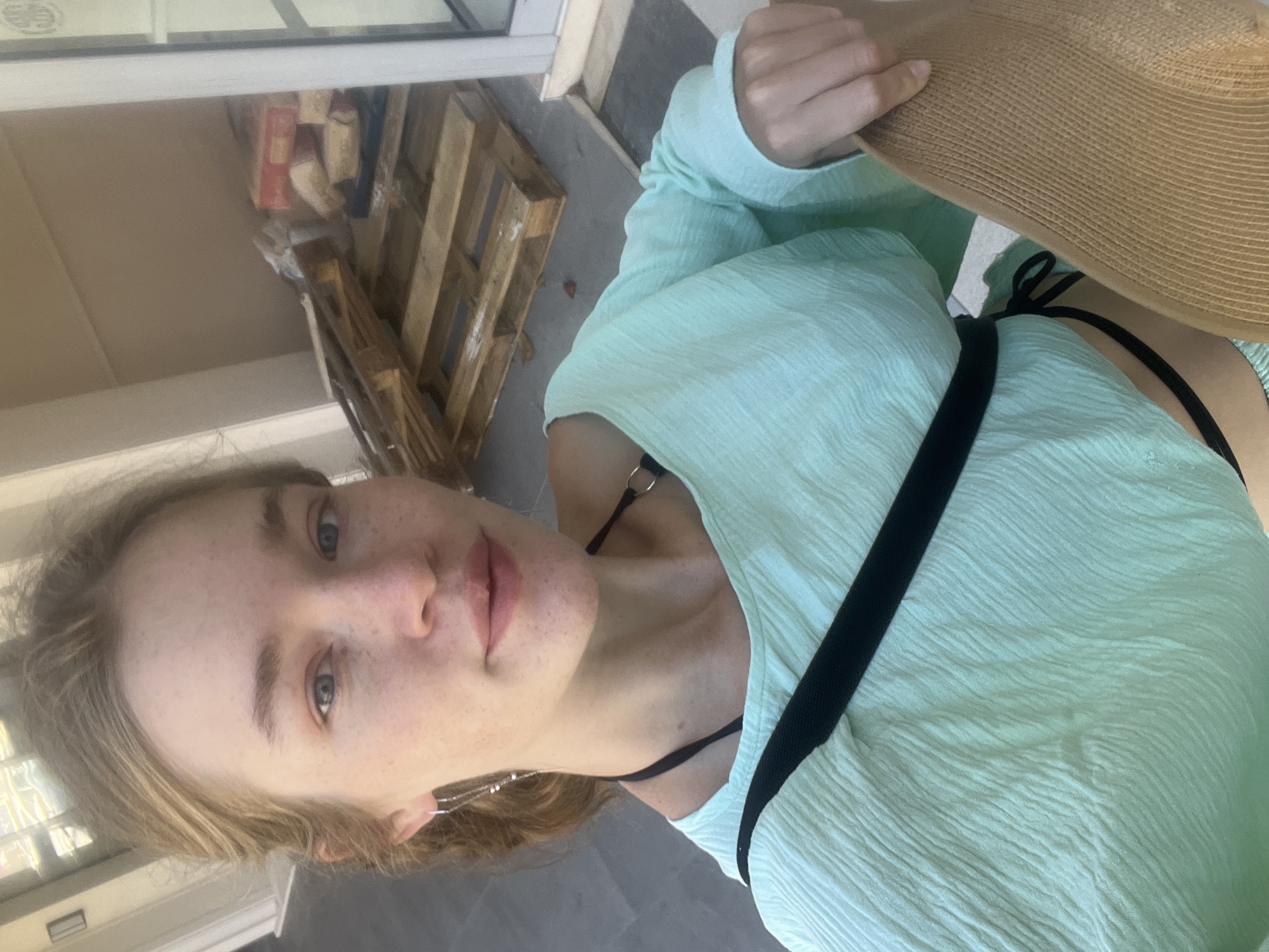 A person, fair-skinned with freckles and blonde hair, is posing for a selfie outdoors. They are wearing a light green, textured top and a black bikini top visible at the neck and strap. A brown straw hat is partially visible, held in their hand. The background includes a wooden pallet and what appears to be a building facade, suggesting an urban location. Given the prompt, the location could potentially be within the city of Budva, Montenegro, although specific landmarks are not visible to confirm.