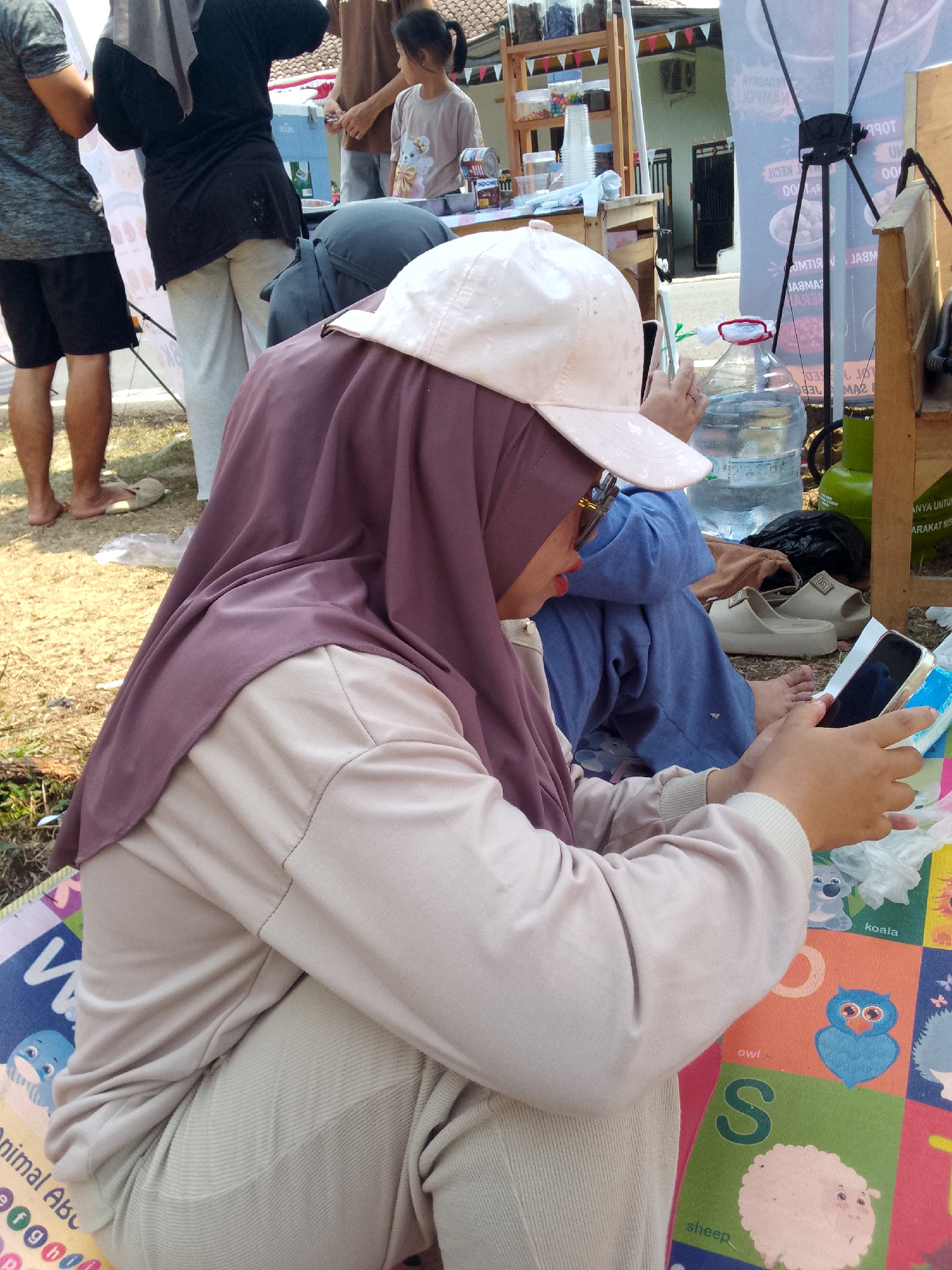 In a street market setting in Mandalawangi, Indonesia, a person wearing a light pink baseball cap and a purple hijab is seated on a colorful mat. They are looking down at a smartphone held in their hands, appearing to be engaged with the device. The person is dressed in a light beige long-sleeved shirt and light gray pants. To their right, a large transparent water jug is visible. In the background, other individuals are present, some standing and one child is looking towards a stall. The ground is covered with dry grass and dirt. There are various items on display and a wooden structure that suggests a market stall.