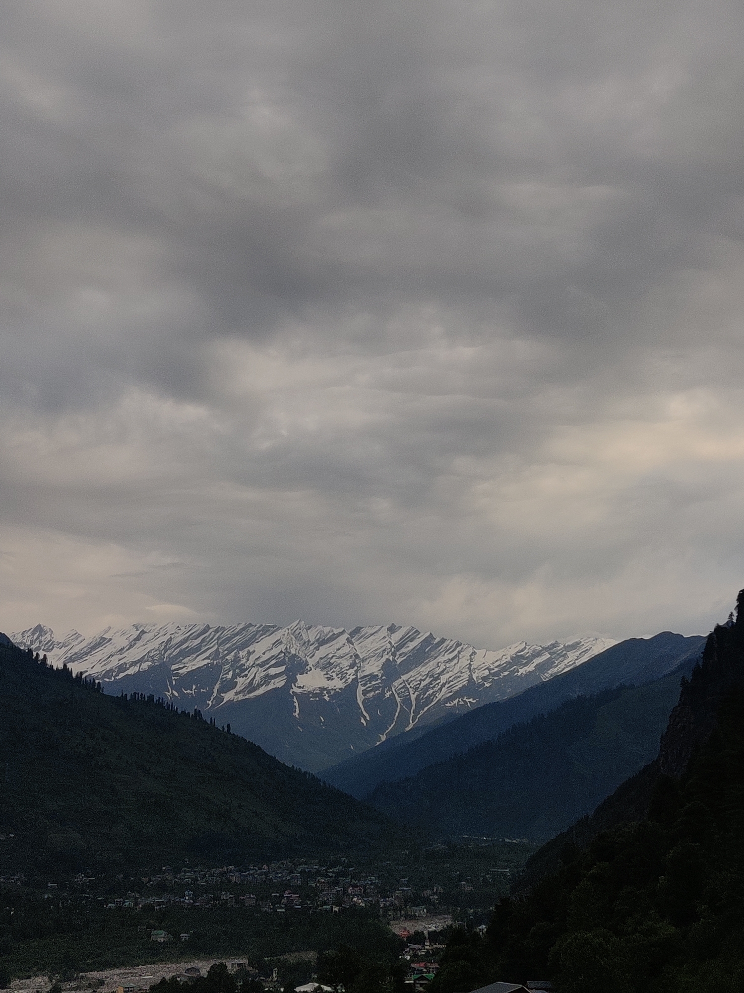 The image depicts a panoramic view of a valley in Vashisht, India, with snow-capped mountains in the background. The sky is overcast with dark, heavy clouds, suggesting impending rain or a cloudy day. The mountains, with their jagged peaks, are covered in a substantial layer of snow, contrasting with the dark green, tree-covered slopes of the lower hills. A small town or village is nestled within the valley, with buildings clustered together. The overall atmosphere is serene and majestic, emphasizing the natural beauty of the Himalayan landscape. The lighting is diffused due to the cloud cover, resulting in a somewhat muted color palette.