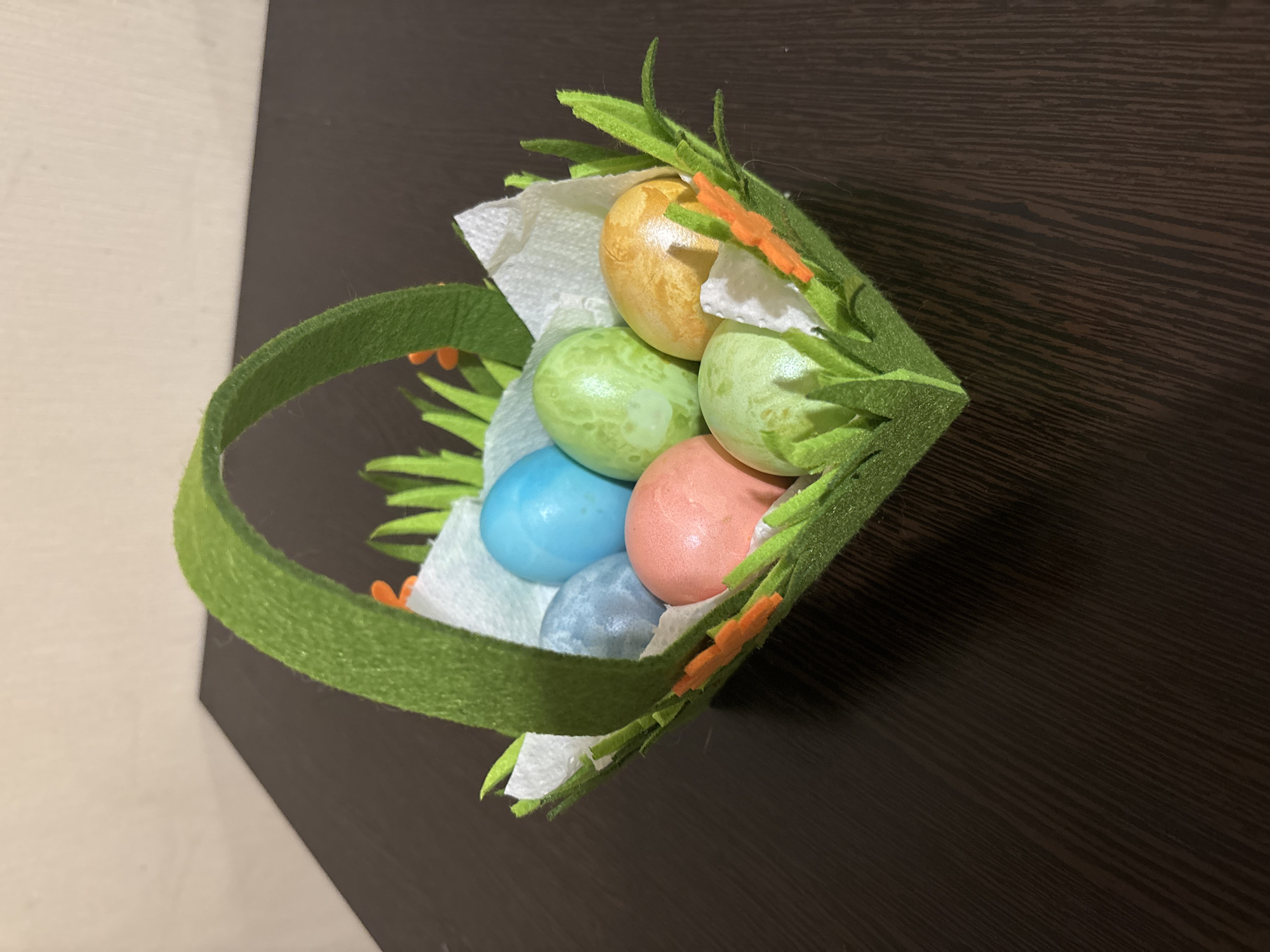 A slightly elevated, medium-angle shot shows an Easter basket filled with six colored eggs, resting on a dark brown wooden surface. The basket is constructed of green felt, with additional green felt cut into leaf shapes surrounding the basket's edge. Small orange felt embellishments are also attached to the basket's leaves. The handle of the basket, also made of green felt, is on the left side of the frame. Inside the basket, crumpled white tissue paper provides a nest for the eggs. The eggs are colored in varying shades of yellow, green, blue, pink, and grey.
The location is not provided in the image or the context.