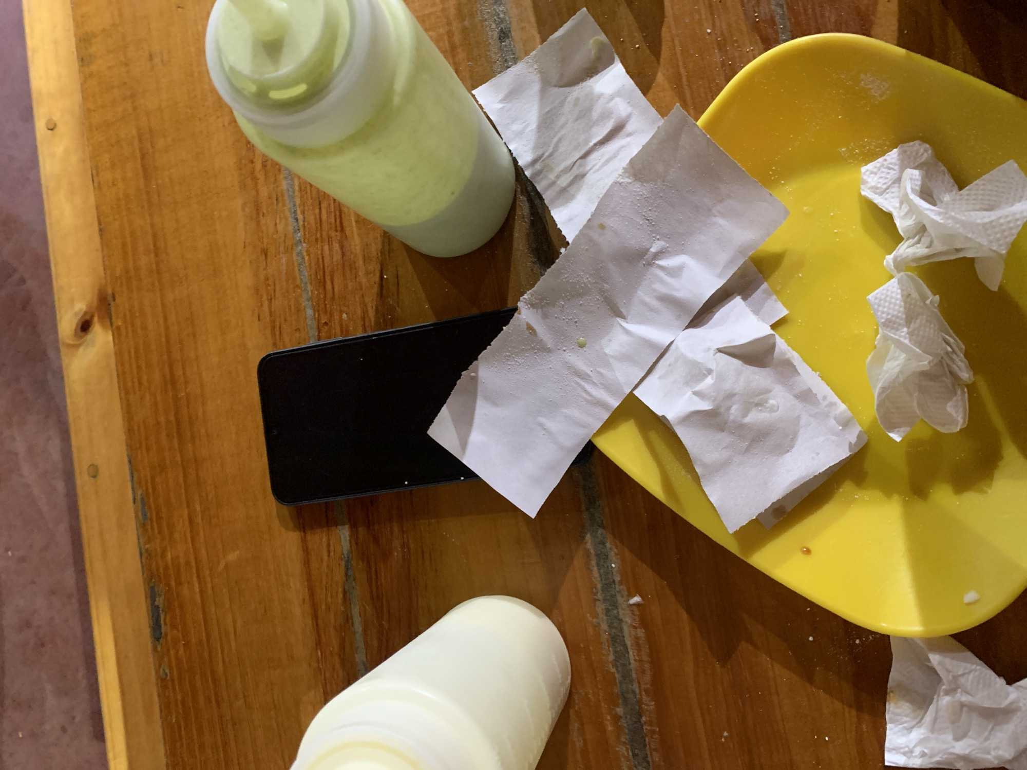 The image provides an overhead, close-up view of a rustic wooden table after a meal, likely in Santa Ana, Venezuela. The light brown wooden surface exhibits prominent grain and some darker marks.
In the upper left quadrant, a translucent squeeze bottle containing a pale green liquid, presumably a condiment or sauce, stands upright. Another off-white, opaque squeeze bottle lies partially visible in the lower left, also appearing to be a condiment container. A black smartphone is centrally placed on the table, with its screen facing downwards or turned off.
To the right, a bright yellow plastic plate holds several crumpled, used white paper napkins. Additional soiled white napkins, showing small food particles or crumbs, are scattered directly on the table surface. The yellow plate also has some light residue, further indicating it was recently used for food. The scene conveys the casual aftermath of eating, with no people visible and no discernable text on any of the objects. The lighting suggests daytime.