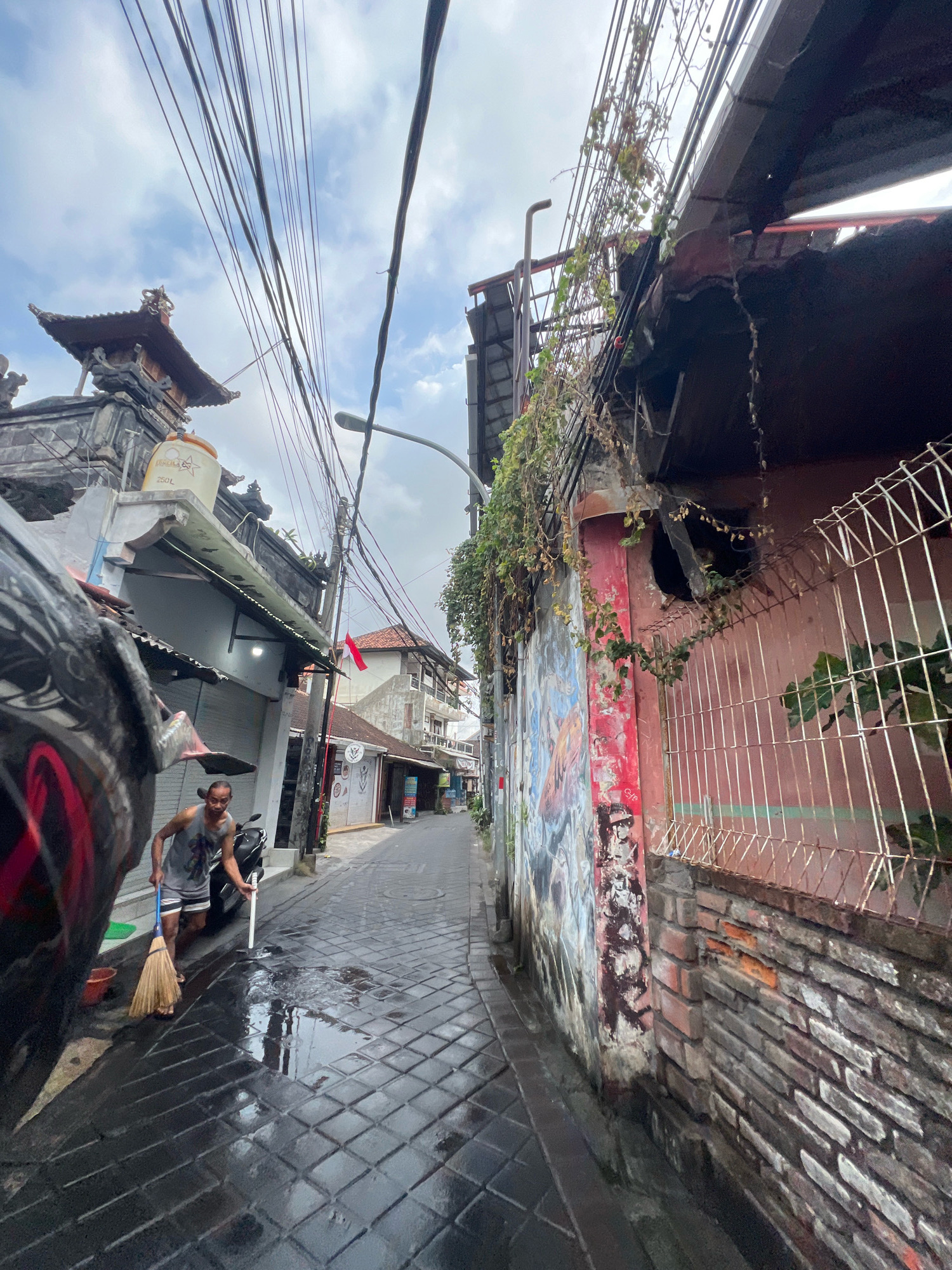 In Kuta, Indonesia, a man in a grey tank top and patterned shorts sweeps a wet, paved alleyway with a broom. The alley is lined with buildings displaying a mix of traditional Balinese architecture and modern facades. Overhead, a dense network of electrical wires crisscrosses the sky. One building features intricate stone carvings and a large water tank. Another wall is adorned with colorful graffiti. A metal fence with a diamond pattern encloses a section of the property. A motorcycle is parked to the man's right. The ground is wet, suggesting recent rain or cleaning. The sky is overcast with scattered clouds.