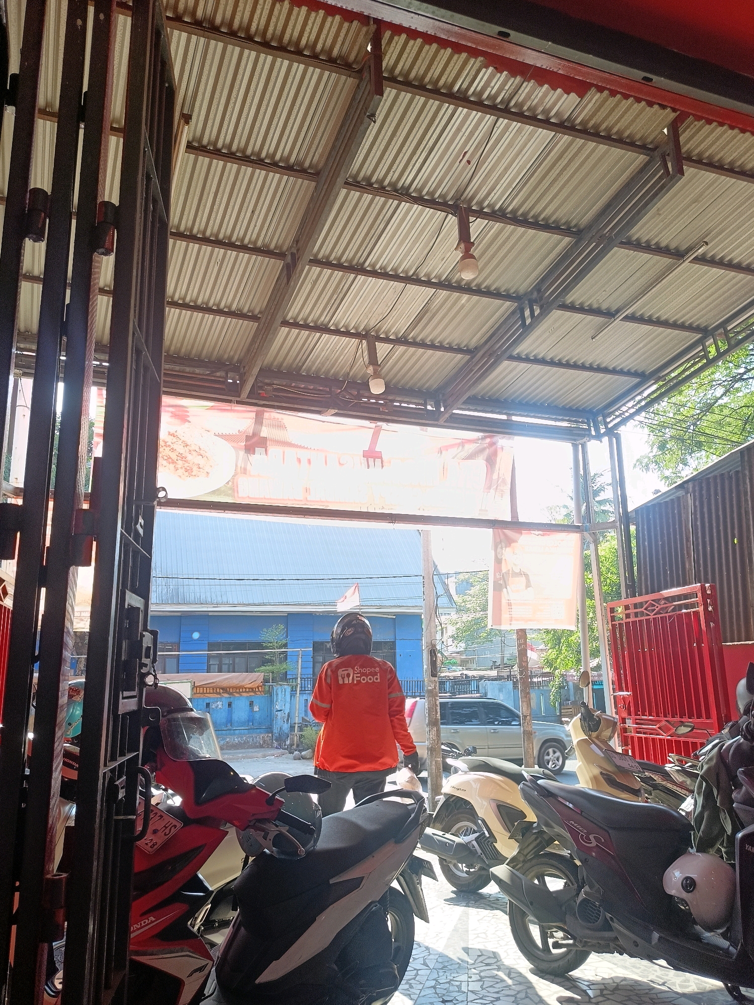 A delivery driver wearing an orange Shopee Food jacket stands in the foreground under a corrugated metal roof. The driver is facing away from the camera, looking out towards a street scene. Multiple parked motorcycles are visible in front of the driver, some with helmets on them. In the background, a blue building and trees are visible. The scene appears to be outdoors, possibly a street-level establishment or an alleyway. The lighting suggests it is daytime.
