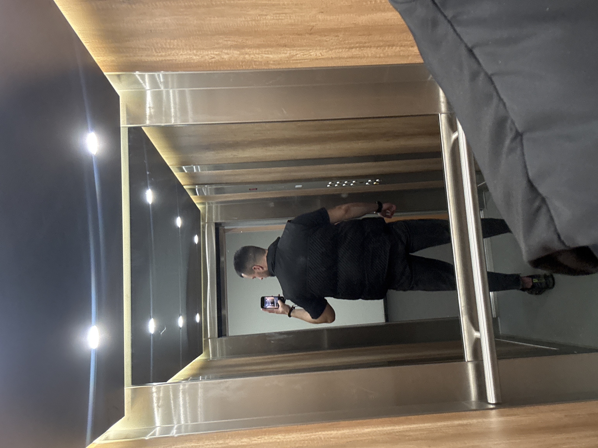 A person in an elevator in Sofia, Bulgaria is taking a photo with a smartphone. The elevator has a mirrored back wall. The elevator's control panel is visible near the top of the mirrored wall. The person is wearing dark clothing and stands near the back of the elevator. A portion of another person is visible in the right corner of the image. The elevator interior is lit with overhead lights. The walls and ceiling of the elevator are finished with wood and metal.