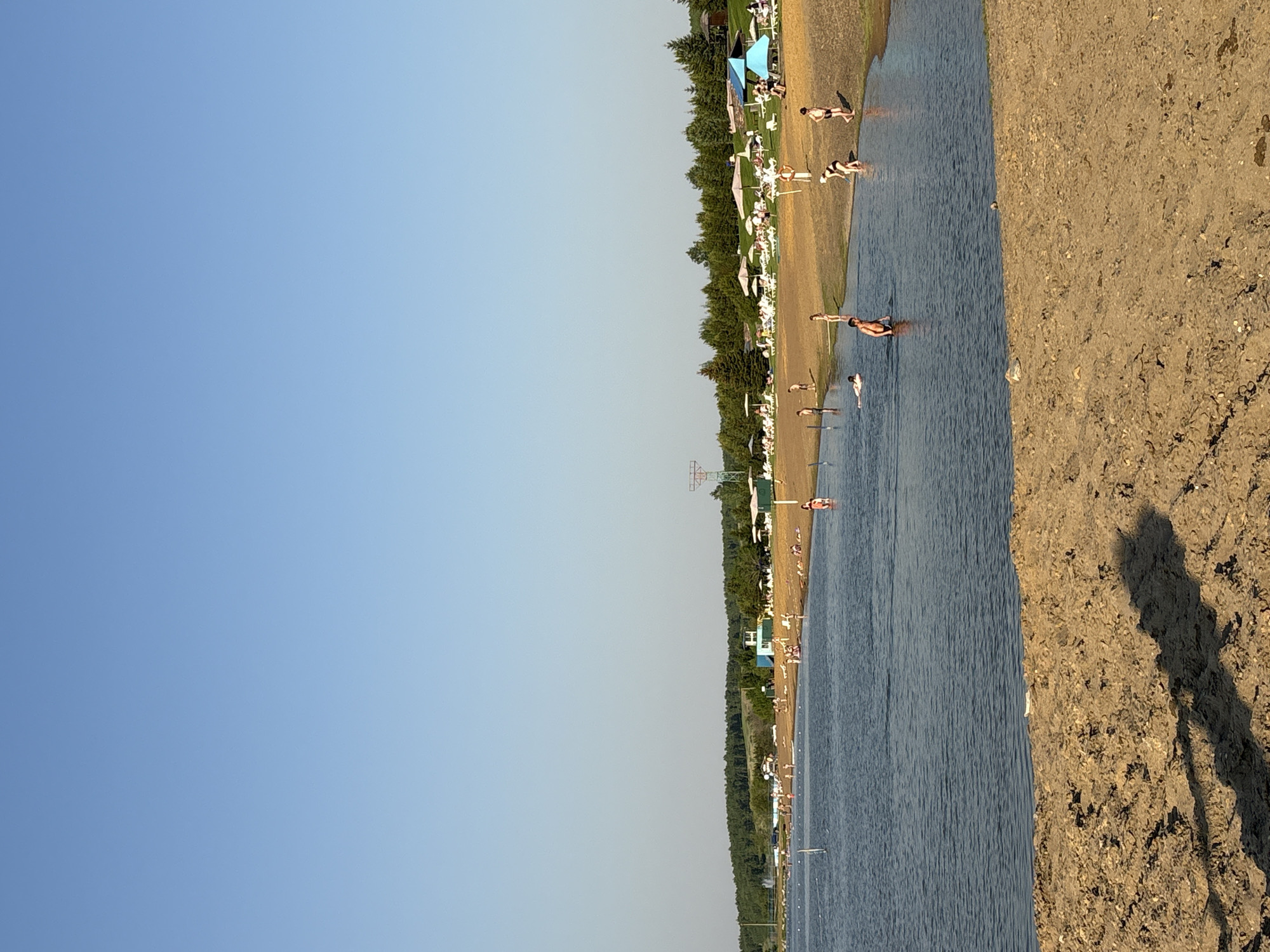The image depicts a leisure scene on a sunny day at a lakeside beach in Emel'yanovo, Russia. The upper half of the frame is dominated by a clear, bright blue sky with no visible clouds. Below, a calm body of blue-grey water, likely a lake or reservoir, occupies the mid-ground, showing gentle ripples and a few white buoys marking a designated area.
The foreground features a sandy, light-brown beach with visible pebbles and darker, moist patches, where long shadows stretch towards the lower left, indicating a low sun angle. A prominent, elongated shadow extends from the bottom left corner, likely cast by the photographer or an unseen object.
Numerous individuals are engaged in various recreational activities. In the water, at least five people are visible: one person is floating on their back on a white board in the far left, while others wade or stand in the shallows. On the sandy beach closer to the water, several individuals walk or stand, including one person bending over near the water's edge, and another further back holding a long white pole. On the far right of the beach, a small group stands near the water.
Beyond the immediate shoreline, a grassy area hosts an organized leisure setup. White lounge chairs are arranged beneath numerous white and a few blue sun umbrellas and canopies, occupied by multiple individuals relaxing. Further back, a light blue and white building with a distinct observation tower stands amidst the trees, possibly a lifeguard station or resort facility. Other small structures are scattered throughout. The background is defined by a dense line of coniferous and deciduous trees, forming a green forest across distant hills. A tall, reddish-brown metal structure, possibly a pylon or part of a ski lift, is partially visible on the far right behind the trees.