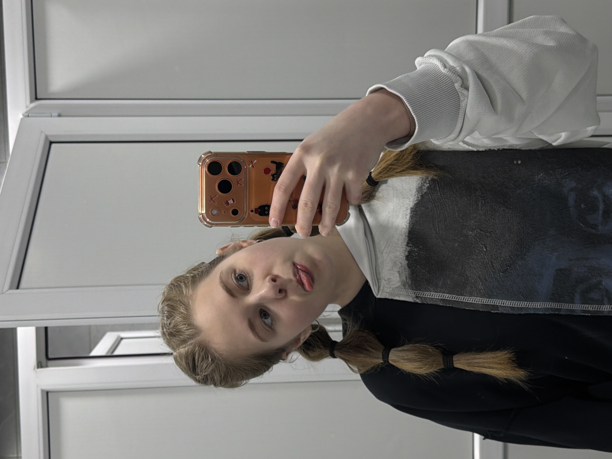 A young woman takes a selfie in front of a window in what appears to be a bathroom or similarly utilitarian room. The woman has fair skin, blue eyes, and blonde hair styled in pigtails secured with black elastics. She is wearing a black and white hoodie. She is holding a brown smartphone with cartoonish decorations on its case. The background consists of a white-framed window and light-colored walls. There is no indication of Perm, Russia, in the image.