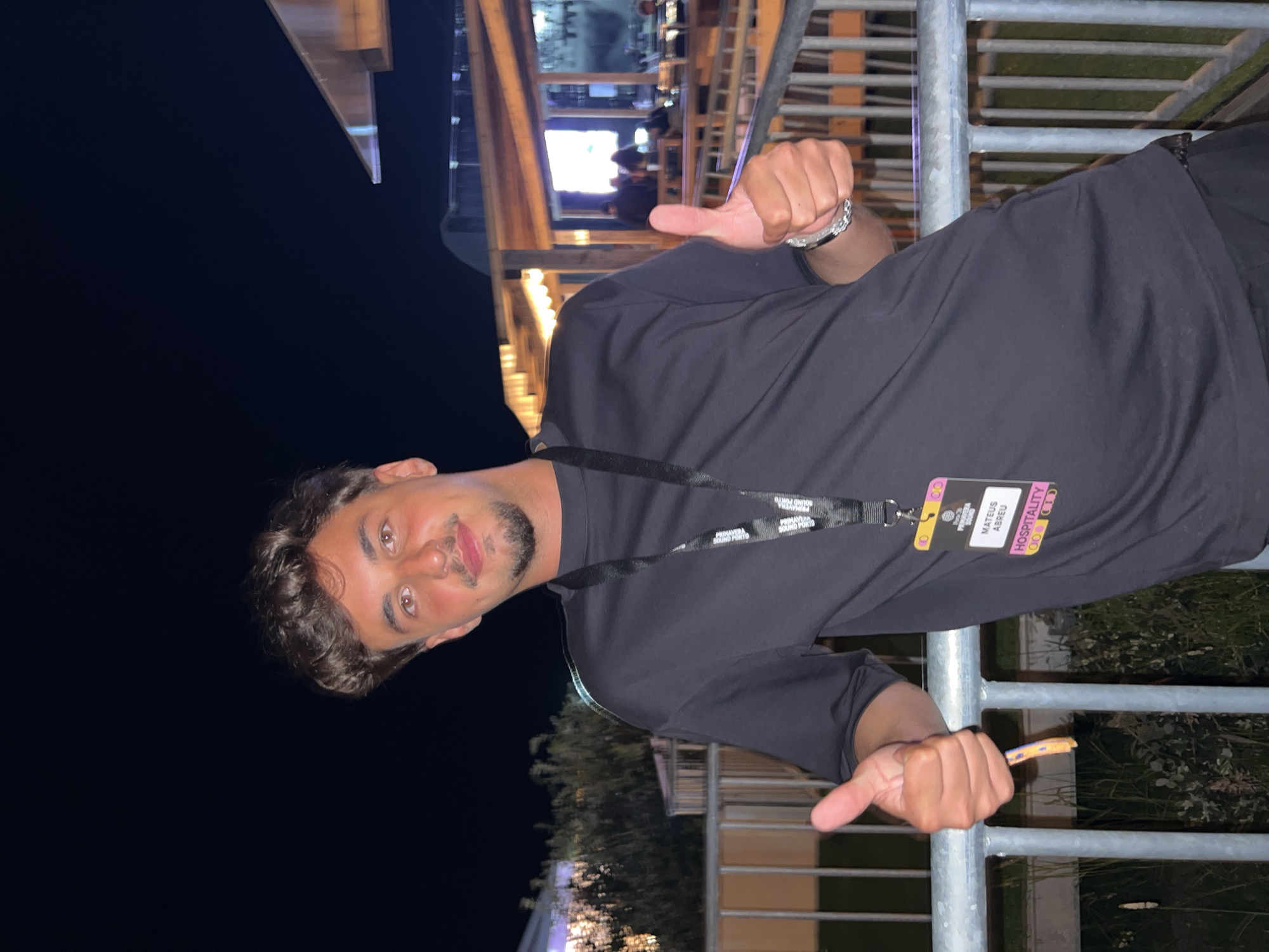 A young man with short, dark, curly hair and a mustache is standing outdoors at night, in front of a metal railing. He is wearing a black t-shirt with a lanyard around his neck. The lanyard has a badge attached to it that reads "MATEUS ABREU HOSPITALITY". He is giving a thumbs up with his left hand, and his right hand is also giving a thumbs up. Behind him, there is a building with lights and people sitting at tables. The environment appears to be a venue for an event, potentially Primavera Sound Porto, given the badge. The background suggests an outdoor area at a festival or concert venue in Porto, Portugal.