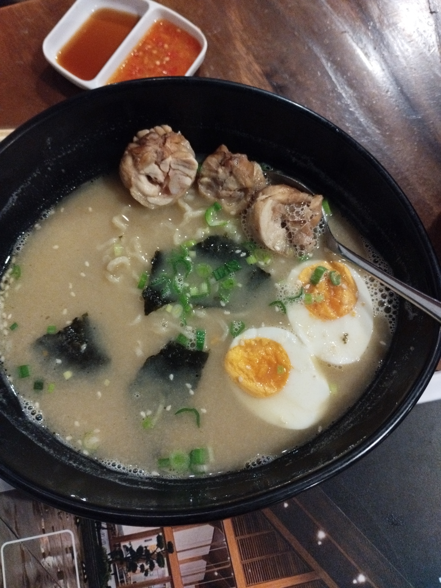 A black bowl of ramen with broth, noodles, chicken, seaweed, green onions, and halved boiled eggs sits on a wooden table. To the left of the bowl is a white dish with two compartments, one containing a reddish sauce and the other a lighter colored sauce. The context of this image has been determined to be Pematang Siantar, Indonesia based on metadata that is not visible in the image.