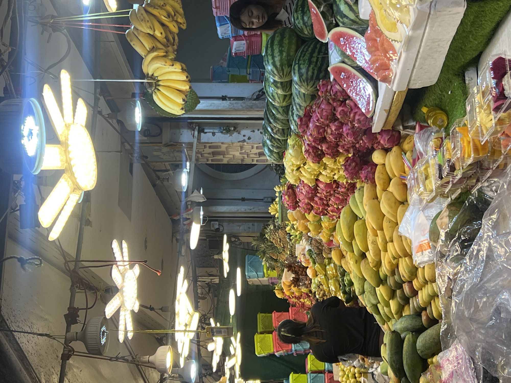 This media file depicts a vibrant fruit stall in a market setting, likely in Karon, Thailand. The scene is filled with an abundance of fresh fruits, including bunches of yellow bananas hanging from above, stacks of watermelons, and piles of dragon fruit in shades of pink and purple. Various other fruits, such as mangoes, pineapples, and possibly lychees, are also visible.
Several decorative, star-shaped lights are strung overhead, illuminating the market. In the background, a person is visible, appearing to be working at the stall or a customer. The overall atmosphere suggests a bustling marketplace. The lighting and the variety of fresh produce indicate it is likely daytime, with the market in full swing. The focus is on the colorful and abundant display of fruits.