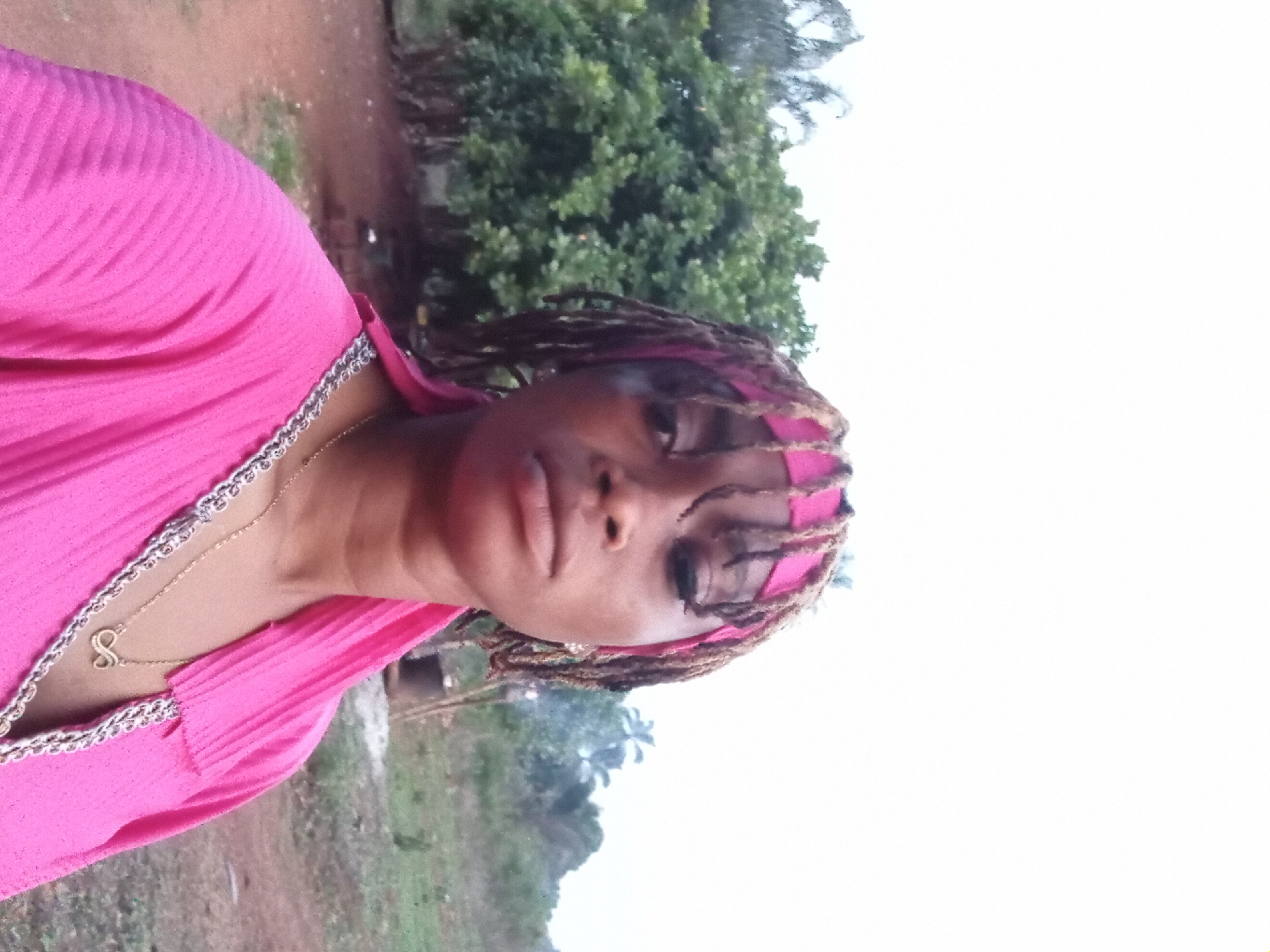 A young woman is depicted in a portrait-style photograph, captured outdoors in what appears to be Ogbe, Nigeria. She is positioned on the left side of the frame, gazing directly at the camera with a neutral expression.
She is wearing a bright pink garment, possibly a top or dress, featuring a textured, pleated fabric and a decorative silver-toned trim along the neckline. A delicate gold chain necklace with a small, gold pendant, resembling a stylized letter 'S' or an infinity symbol, hangs around her neck. Her hair, styled in dreadlocks or braids, is partially covered by a pink band or scarf worn across her forehead.
The background features lush green trees and dense foliage, suggesting a natural or semi-natural environment. The ground closer to the subject appears to be unpaved earth. A significant portion of the right side of the background is dominated by a bright, almost white sky, indicating it is daytime with strong sunlight or overcast conditions. The overall activity appears to be posing for a casual outdoor photograph. There is no visible text present in the image.