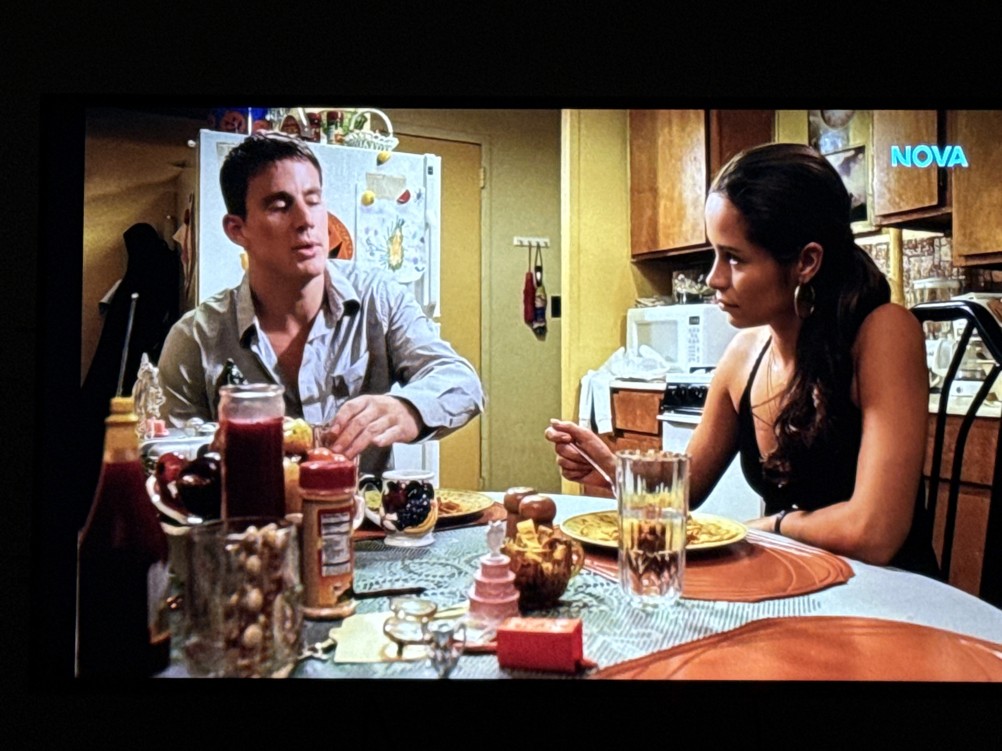 In a dimly lit kitchen, a man and a woman are seated at a table, engaged in conversation and eating. The man, with short brown hair, wears a light-colored, button-down shirt. He is holding a fork and a knife, and appears to be in mid-sentence. The woman, with long dark hair pulled back, is wearing a black tank top and hoop earrings. She is looking away from the camera, with a fork in her hand, seemingly about to eat. The table is set with plates of food, glasses, and various condiments. The kitchen environment appears dated, with wooden cabinets and a microwave visible in the background. A refrigerator stands against the wall, adorned with various magnets and papers. The overall atmosphere suggests a casual, intimate moment between the two individuals. There is no visual information in the image to suggest the location is in Delmenhorst, Germany.