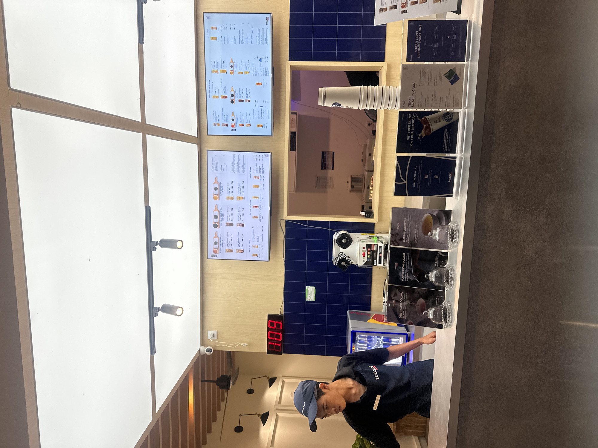 The image displays the interior of a bubble tea shop, focusing on the counter and service area. A male employee, wearing a dark blue cap and a dark blue uniform shirt with the "teazzi" logo, is positioned behind a clean, light-colored counter, looking downwards.
Above the counter, two large digital menu screens prominently display various drink options, including "SIGNATURE SELECTION," "FOUR SEASONS OOLONG TEA," "DEEP ROAST OOLONG TEA," "JASMINE GREEN TEA," "FRUIT TEA SERIES," and "HONEY SERIES," along with corresponding prices and images. To the right of the screens, the wall is accented with dark blue tiles.
On the counter, several acrylic display holders feature promotional and informational cards. These include a "TEAZZI LOYALTY CARD," an offer to "GET FREE DRINK ON YOUR BIRTHDAY," "SUGAR LEVEL RECOMMENDATIONS," and a "KEBIJAKAN HALAL" (Halal Policy), indicating a location where such a policy is relevant, consistent with the given context of Kuta, Indonesia. A stack of white paper cups is also visible, along with small glass domes, possibly for displaying samples.
A red digital clock on the tiled wall shows "1:08," likely signifying 1:08 PM. Below the clock, a portion of a Red Bull refrigerator can be seen. The setting appears modern and well-maintained, with bright ceiling lights and wooden paneling contributing to a clean aesthetic. A security camera is mounted on the wall. The employee seems to be engaged in routine work, suggesting the shop is open and operational.