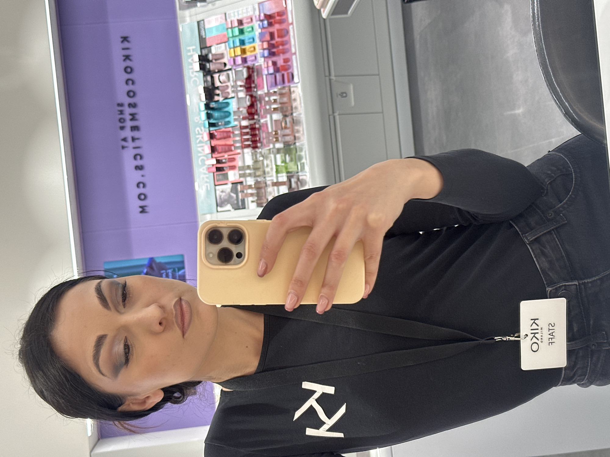 A woman is taking a selfie in what appears to be a Kiko Cosmetics store. The store is located in an unknown city. She is wearing a black shirt with the Kiko logo and a name tag indicating she is a Kiko staff member. Behind her, there's a display of makeup products. The store is well-lit and appears modern.