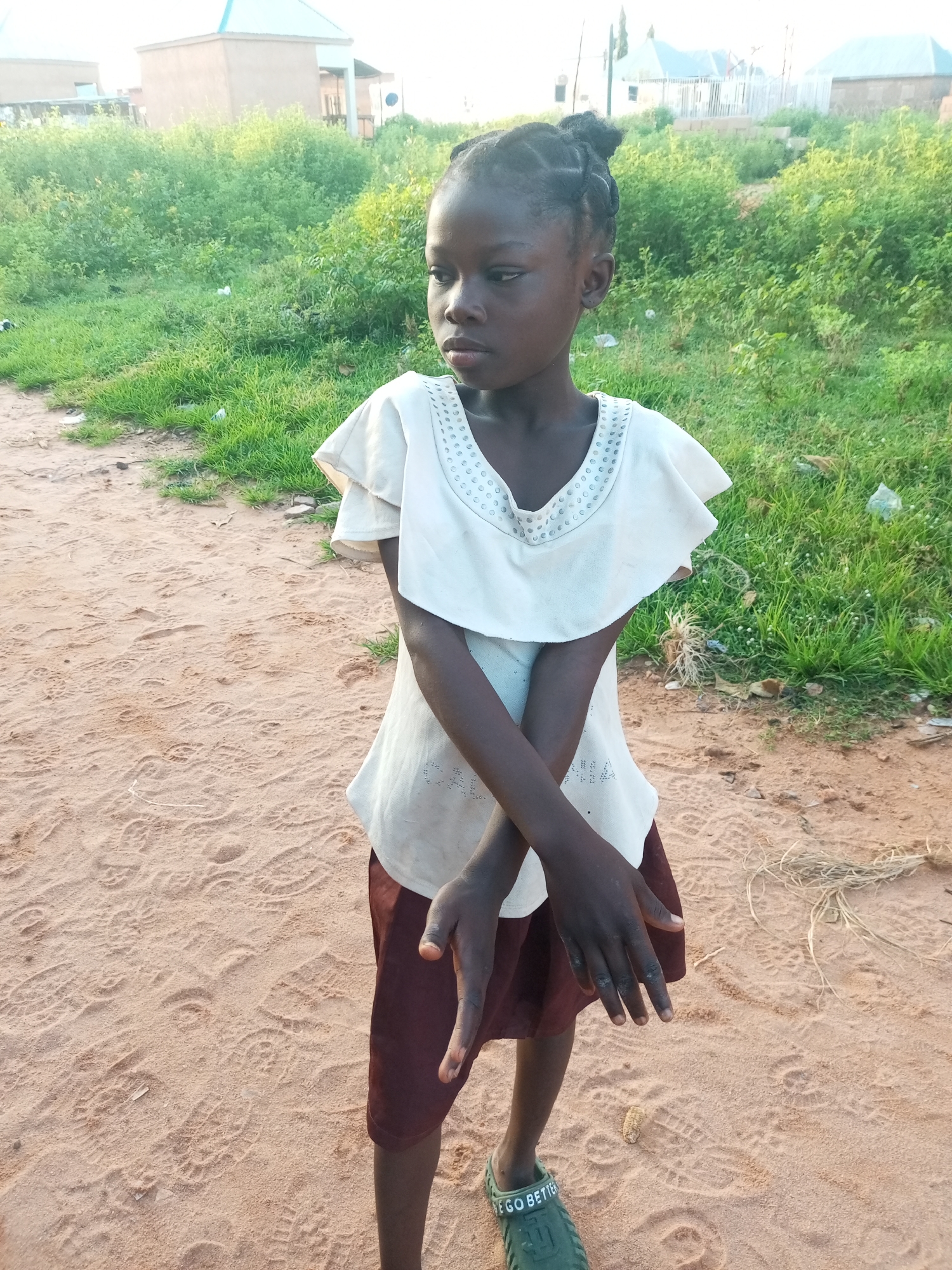 A young girl stands in the dirt outside in Tempure, Nigeria. The girl is facing left, with her arms crossed. She has braided hair pulled up into a bun. She is wearing a light-colored shirt with a ruffled collar and a maroon skirt. Her feet are bare, except for a green sandal on her left foot. The background is filled with green brush and buildings. The ground is sandy, with many footprints visible.