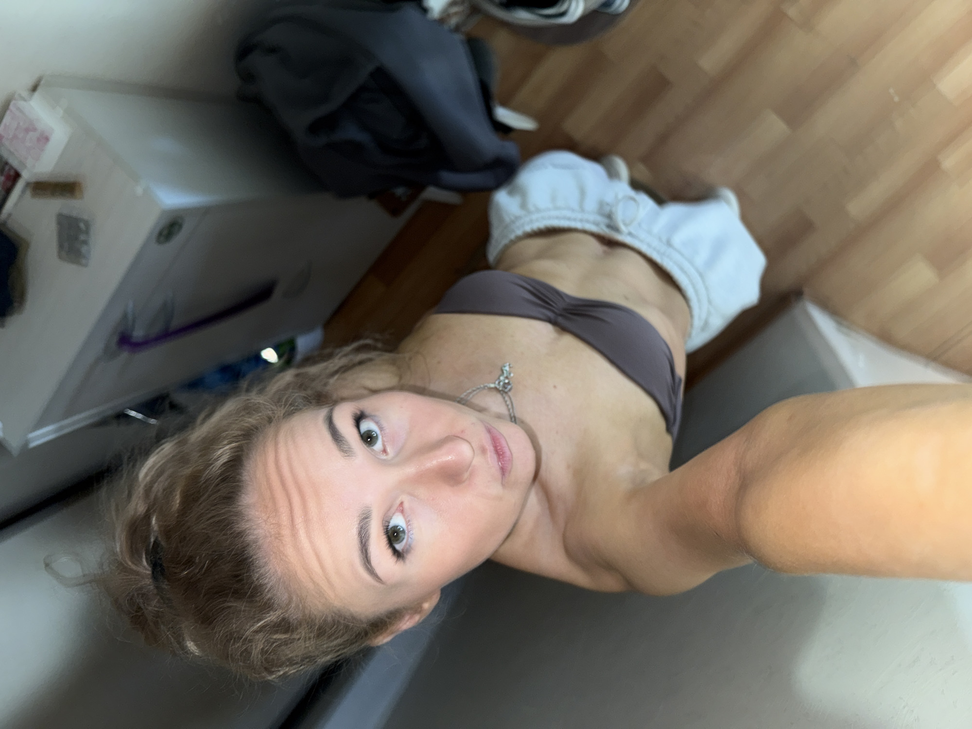 A selfie taken in an indoor location, possibly a kitchen or laundry area, with a woman as the primary subject. The woman is fair-skinned, with curly blonde hair, green eyes, and is wearing a brown bra top and light-colored sweatpants that are pulled down. She is also wearing a silver necklace. The background includes a white cabinet, a pile of clothing (gray and white), and flooring that appears to be wood laminate. The image is taken from a low angle, looking upwards at the woman. The city of Chaikovsky, Russia, is not discernible from the details provided in the image.