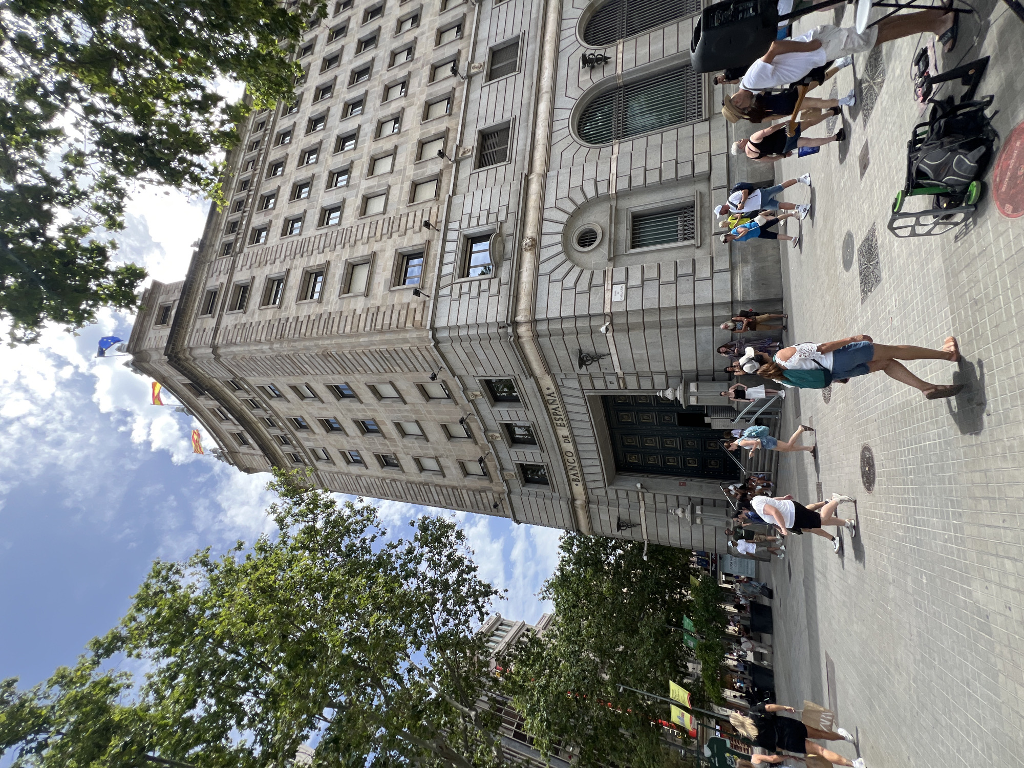 The image depicts a bustling street scene in Barcelona, Spain. A grand, ornate building, identified by the text "BANCO DE ESPAÑA" above its entrance, dominates the background. The architecture is neoclassical, characterized by its stone facade, numerous windows, and decorative elements. Flanking the building are leafy green trees, casting dappled shadows on the tiled pedestrian walkway.
Several people are visible in the foreground and middle ground, walking and milling about. They are dressed in casual summer attire, suggesting a warm, sunny day. Some individuals are captured mid-stride, while others appear to be gathered, possibly socializing or observing. The mood seems relaxed and leisurely, indicative of a typical urban environment on a pleasant day.
A flag, possibly the flag of Spain or Catalonia, is visible flying from a pole on the building, adding a touch of national or regional identity to the scene. The lighting suggests it is daytime, likely late morning or early afternoon, given the bright sunlight and clear blue sky with scattered clouds. The presence of multiple people and the general activity imply this is a public space, likely a well-trafficked area in the city center.