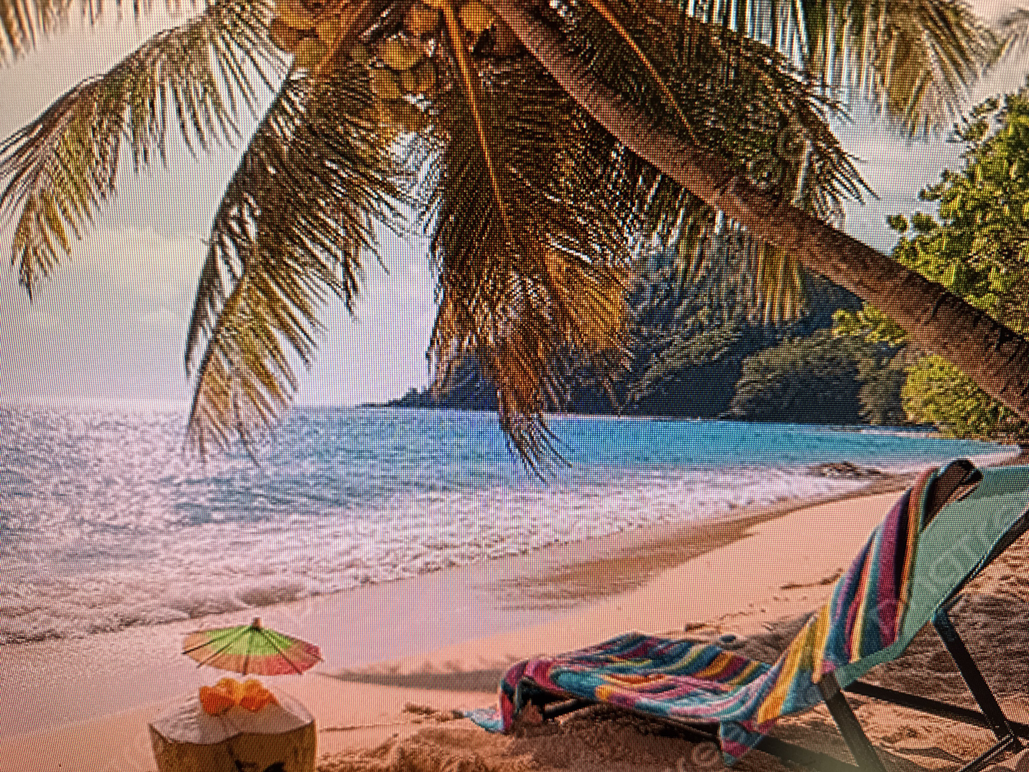 This media file depicts a serene tropical beach scene. The foreground is dominated by a sandy beach with a few select objects. A striped beach chair, draped with a colorful towel, sits prominently on the right side of the frame. To the left, a coconut with a small, colorful umbrella and what appear to be orange slices rests on the sand.
The middle ground features the gentle lapping of waves on the shore, with the ocean stretching out to the horizon. The water is a vibrant blue, indicative of clear tropical waters. In the background, lush green foliage and trees line the coastline, suggesting a verdant, island setting. A portion of a palm tree trunk and its fronds are visible in the upper part of the image, casting shadows on the scene and reinforcing the tropical theme.
The lighting suggests it is daytime, with bright sunlight illuminating the beach and sea. The overall mood is one of relaxation and tranquility, evoking a sense of vacation and escape. There are no people visible in the frame, emphasizing the peaceful solitude of the location. No text is present in the image. Despite the location context provided as Szczecin, Poland, the visual elements strongly suggest a tropical environment, not a Polish location.