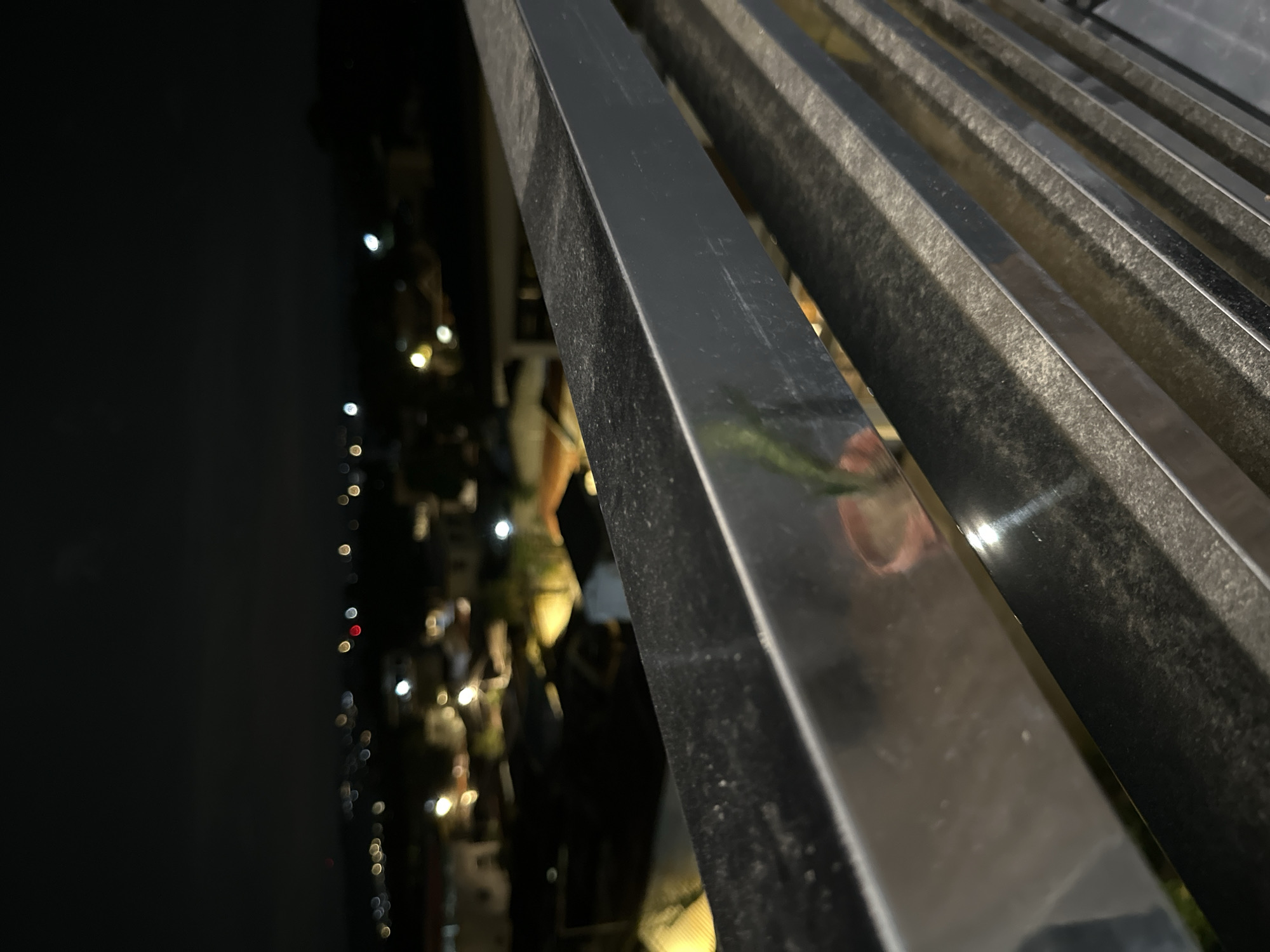 The image shows a close-up view of metal railings in Davao City, Philippines. The railings are dark and appear to be slightly reflective. Behind the railings, there are blurry lights suggesting a cityscape at night. People are visible in the background but are indistinct due to the low light and depth of field. The scene is dark, with the railings being the primary focus.