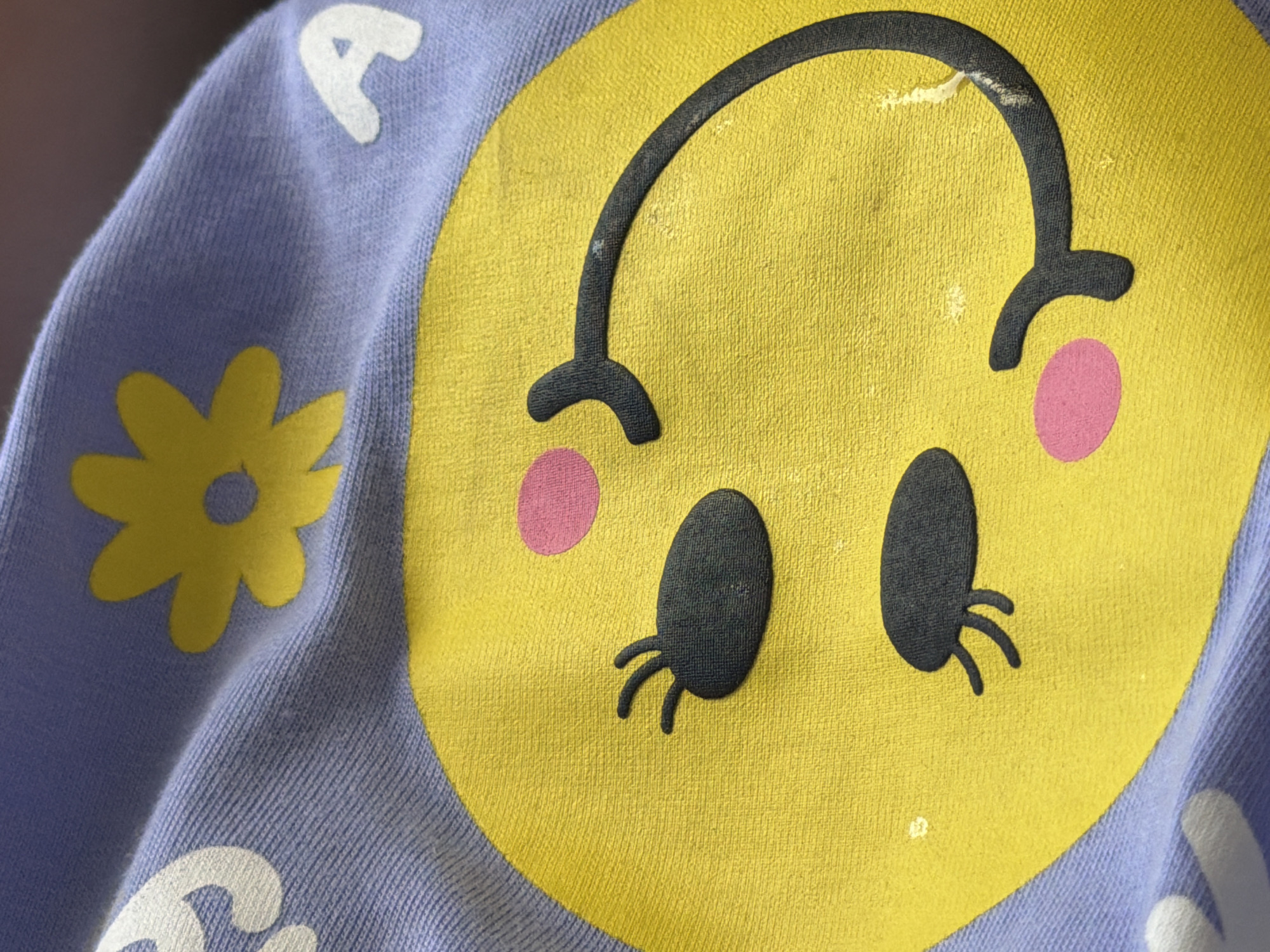 A close-up, high-angle shot focuses on a light purple fabric with a yellow circular graphic and other embellishments. The yellow graphic depicts a stylized smiley face with closed eyes, indicated by two black, oval shapes with three short lines extending from them, resembling eyelashes. Pink circles are placed on either side of the smiley face, representing cheeks. A yellow flower graphic with a dark center is positioned above the smiley face. White, stylized floral or abstract shapes are visible on the left and right edges of the fabric, as well as the letter "A" in white on the upper right. The fabric appears to be a soft, textured material, possibly a child's garment. The background is out of focus and dark, suggesting artificial lighting. The image is taken in the city of Butovo, Russia.