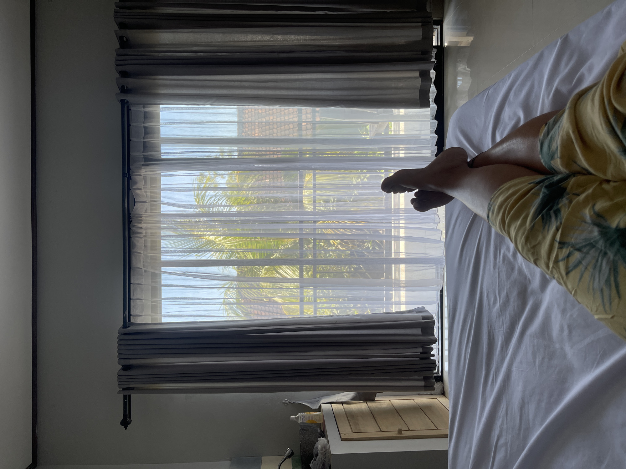 A first-person perspective shot shows a person lying on a white bed with their legs and feet extended towards a large window. The person is wearing yellow shorts with a palm leaf pattern. The window is covered by sheer white curtains and dark gray drapes, through which palm trees and a tiled roof are visible. To the left of the window, a wooden cabinet is partially visible with a white bottle on top. The room appears to be a bedroom, possibly in a tropical location, given the view of palm trees.