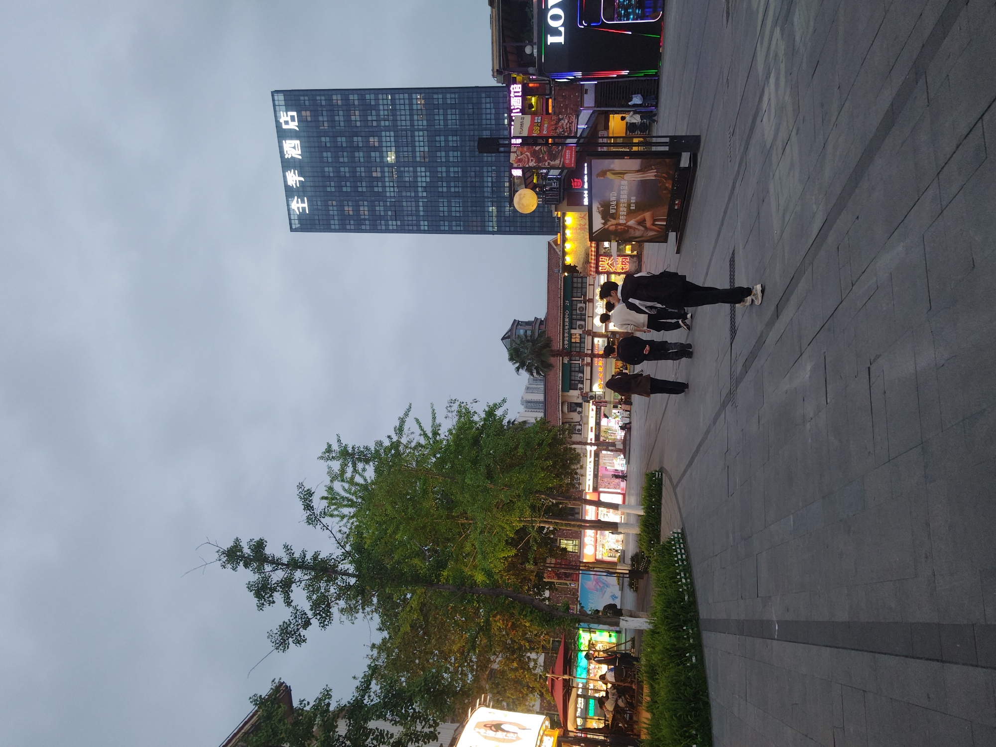 This image captures a vibrant urban street scene in the Chongqing High-tech Zone, China, at dusk or early evening. The sky, a uniform light grey, suggests overcast weather.
In the left half of the frame, a tall, modern glass-fronted building dominates, with the illuminated Chinese characters "全季酒店" (All Seasons Hotel) clearly visible on its facade. Below and to the right of this building, a bustling commercial area unfolds, characterized by numerous storefronts adorned with glowing signs and displays. A prominent large, yellow, spherical light fixture resembling a full moon hangs above the street level, adding a whimsical touch to the urban landscape.
Several people are walking on the wide, paved pedestrian area that occupies the foreground and mid-ground. They appear casually dressed, and their activity primarily consists of strolling through the public space. Along the right edge of the paved path, a narrow strip of green ground cover and small bushes adds a natural element to the otherwise concrete environment. Further left, several mature trees with green foliage provide shade and contrast to the surrounding architecture.
Visible text on signs and billboards includes "全季酒店" (All Seasons Hotel), "发展中心" (Development Center), "火锅" (Hotpot), "LOV," and a large advertisement for "WD&WD 都市漫游生活服饰品牌" (WD&WD Urban Roaming Lifestyle Apparel Brand). The overall impression is one of a lively commercial district coming alive with artificial lights as daylight fades.