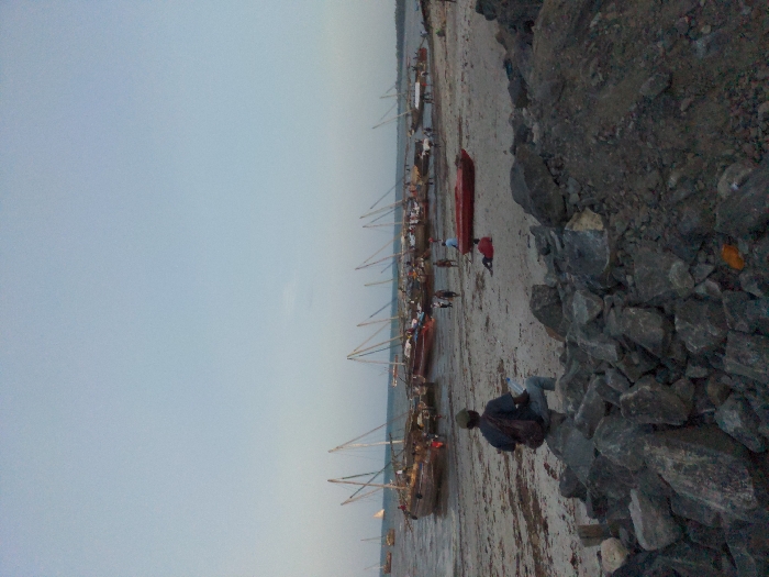 The image captures a coastal scene in Bagamoyo, Tanzania, likely during an overcast day or at dusk/dawn, given the soft, diffused lighting and the muted blue-gray sky. The viewpoint is slightly elevated, looking down upon a beach area.
In the immediate foreground, a rough embankment of dark, irregularly shaped rocks occupies the right side of the frame. A person, wearing a dark jacket and light-colored pants, is seated on these rocks, facing away from the viewer and observing the activity on the shore below.
The midground reveals a wide expanse of sandy or muddy beach, indicative of low tide. A line of numerous traditional wooden fishing boats, possibly dhows, are beached along the waterline. Many of these boats feature tall masts. A distinct red, smaller boat or dinghy is also visible on the sand among the larger vessels. Several other human figures are scattered across the beach, near the boats, appearing to be engaged in various tasks, such as standing, moving, or interacting with the boats. One person near the red boat is notably wearing a red garment.
The background shows the calm waters of the sea or a bay stretching towards a hazy horizon, with a distant landmass faintly visible. The overall scene suggests a bustling fishing village or harbor, depicting the routine activities of preparing or mooring boats. No discernible text is visible within the image. The atmosphere appears tranquil and industrious.